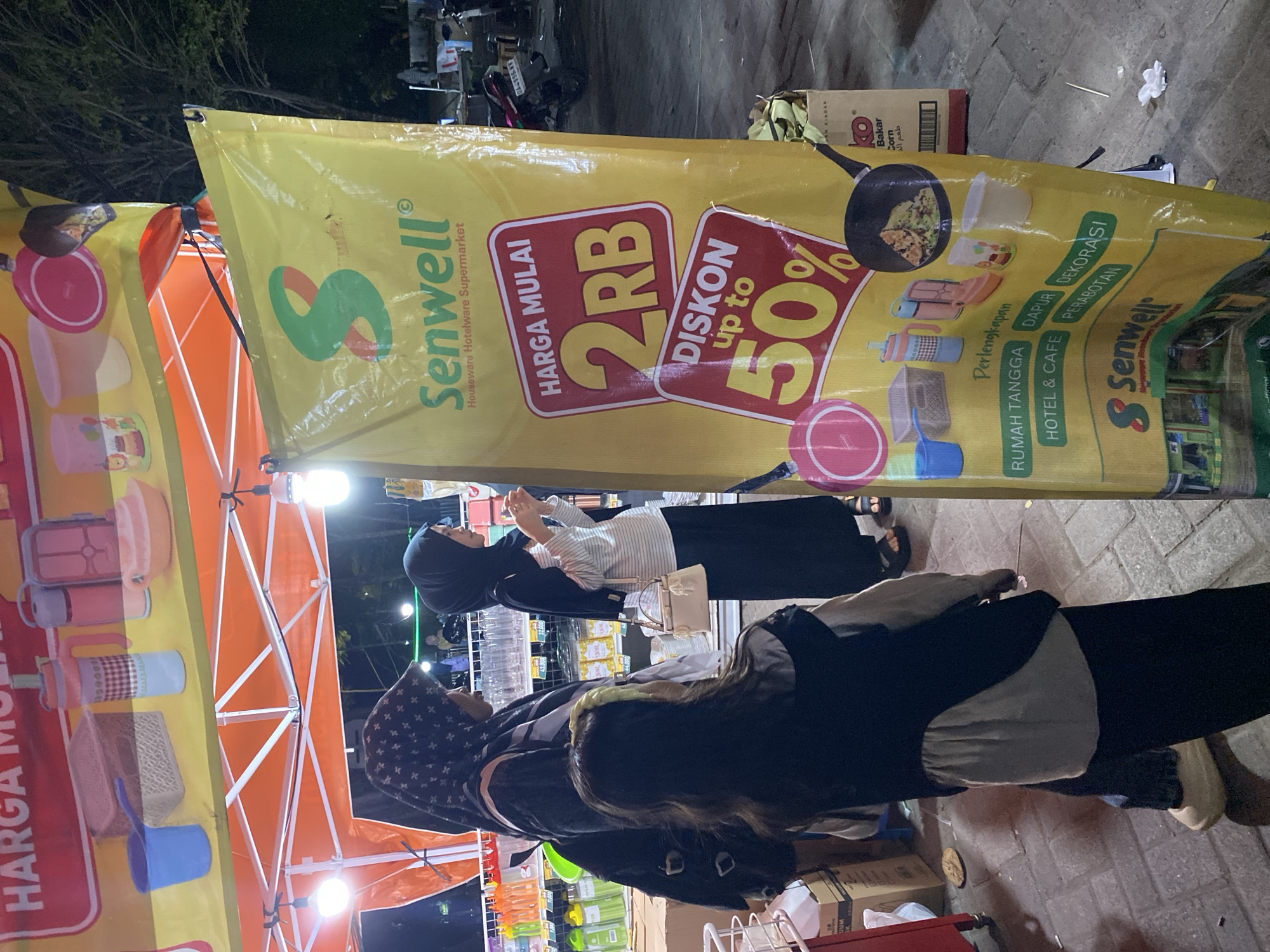 The image depicts a street scene at night in Banjarmasin, Indonesia, likely a market or outdoor shopping area. A large yellow banner for "Senwell Houseware Hotelware Supermarket" dominates the right side of the frame. The banner advertises "HARGA MULAI 2RB" (Prices starting from 2,000 Rupiah) and "DISKON up to 50%" (Discount up to 50%). Below these offers, various houseware items such as containers, cups, and a frying pan with food are illustrated. Text categorizing products includes "RUMAH TANGGA" (Household), "HOTEL & CAFE," "DAPUR" (Kitchen), and "DEKORASI PERABOTAN" (Furniture Decoration).
In the foreground and mid-ground, three individuals are visible. On the left, a woman wearing a dark hijab and a black backpack stands partially obscured. Next to her is a young girl facing away from the camera, wearing a light-colored top under a dark vest and dark pants. To the right of the girl, another woman in a dark hijab and a striped long-sleeved shirt stands holding a small handbag. They appear to be browsing or shopping.
Behind the individuals and the banner, an orange tent structure is set up, illuminated by lights, suggesting a temporary stall or booth. In the background, trees and streetlights are visible, along with glimpses of other stalls and a parked motorcycle with license plate "DA 9556 KX". The ground is paved with cobblestones. The overall atmosphere is that of an evening marketplace.