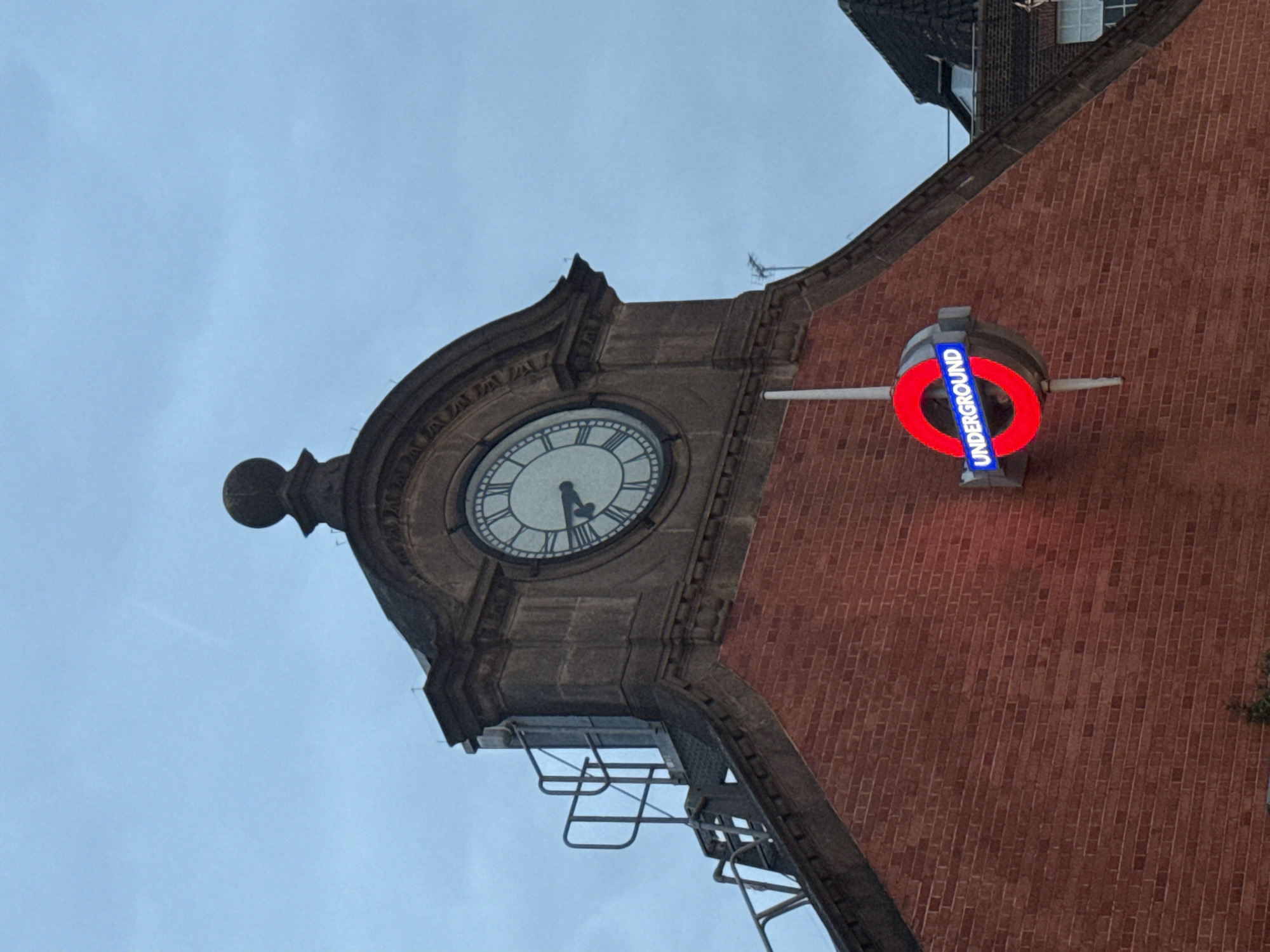 The image shows a London Underground station in the United Kingdom. A clock tower with a white clock face indicates a time of approximately 5:25. An iconic London Underground roundel sign with a red circle and a blue bar displays "UNDERGROUND" on the side of the brick building. A metal staircase extends from the clock tower. The sky is overcast.