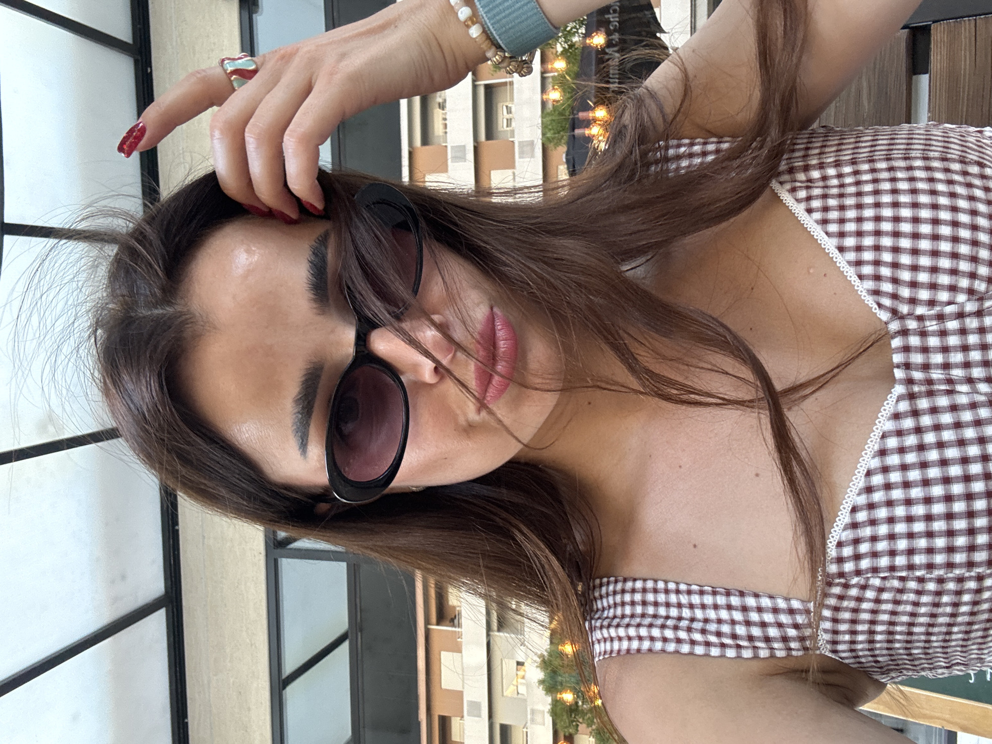In Podgorica, Montenegro, a young woman is captured in a close-up, selfie-style photograph. She is wearing dark, oval-shaped sunglasses that obscure her eyes and a red and white checkered top with a sweetheart neckline. Her long, brown hair is windswept across her face, partially obscuring her features. Her right hand is raised, with a red-painted nail, touching her hair. She is wearing a ring on her right index finger with a turquoise stone and a red accent. On her left wrist, she has several bracelets, including one with light-colored beads and a teal band. The background is softly blurred, showing a building with multiple windows and some outdoor lighting fixtures. The image appears to be taken outdoors, possibly at a cafe or restaurant, given the background elements.