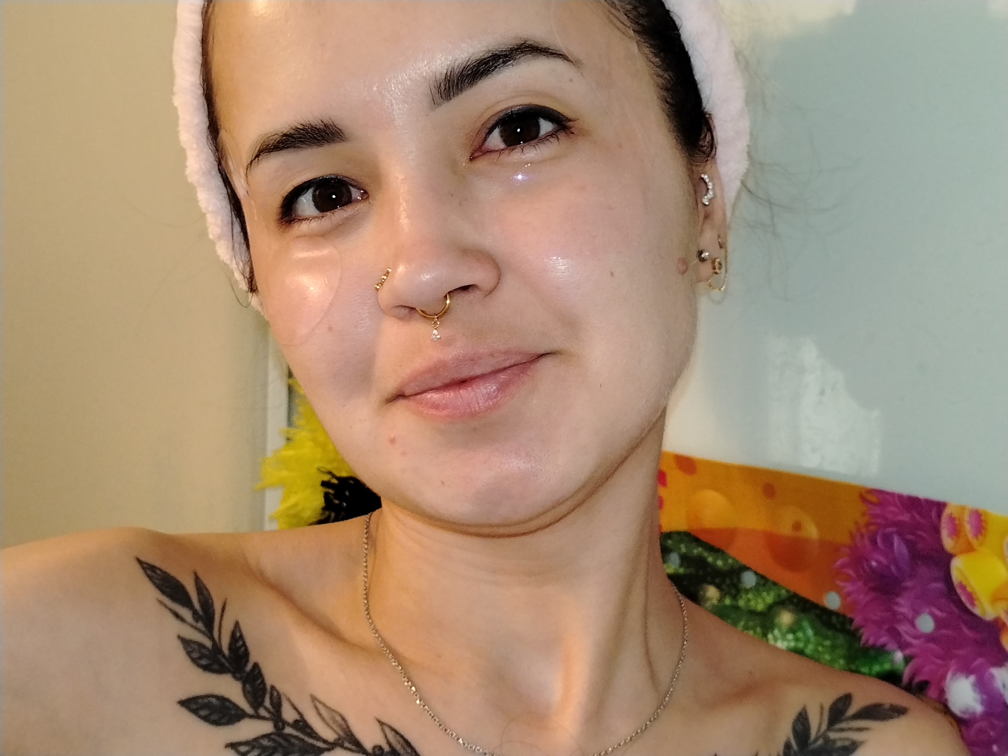 A woman is posing for a selfie. She has dark hair, brown eyes, and fair skin. She is wearing a light pink headband and has several piercings in her ears and nose. Eye masks are under her eyes. A delicate silver necklace is around her neck. A tattoo of leaves is on her left shoulder and chest. A yellow flower is partially visible behind her on the left. A colorful floral blanket is behind her on the right. The environment appears to be indoors. The location is not evident from the image.
