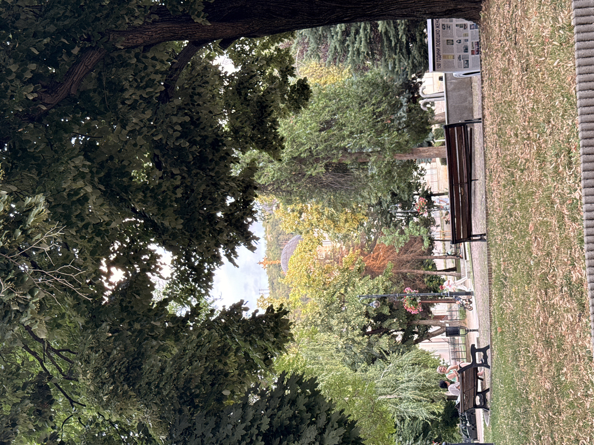 The image captures a park setting, likely in Lovech, Bulgaria, with a prominent view of the Assumption of the Virgin Mary Cathedral on the hillside. In the foreground, two wooden benches are situated on a paved path bordering a grassy area covered in fallen leaves. To the left, two individuals are seated on one bench, engaged in conversation. Further in the background, a person is seen pushing a child on a tricycle, and another individual is visible near a lamppost adorned with pink flowers. Several trees, including deciduous and coniferous varieties, fill the middle ground, with the cathedral's dome and golden cross visible above the trees. A display board with text in Bulgarian is present on the right side of the image, near a large tree. The overall scene depicts a peaceful public space with leisurely activities taking place.