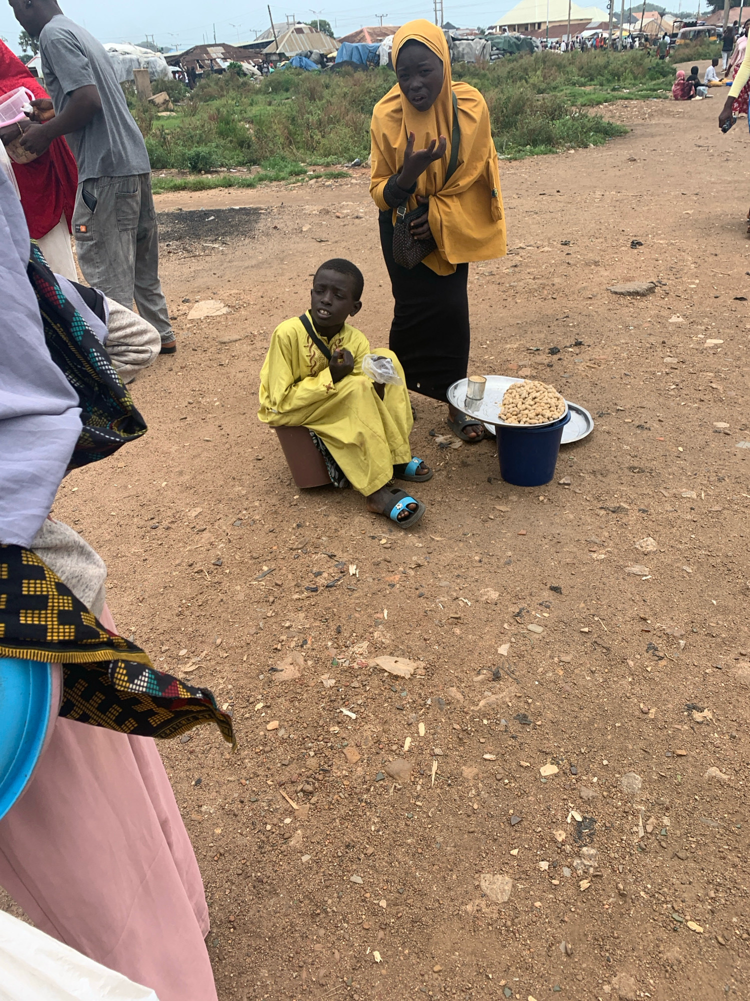 In Bukuru, Nigeria, on a dusty, unpaved road, a young boy wearing a yellow garment sits on a brown bucket, selling peanuts. He is positioned in front of a blue bucket filled with peanuts and a silver tray holding more peanuts and a small tin. To the right, a woman in a yellow hijab and black dress stands looking towards the camera, with her hand slightly raised. To the left, a person wearing a grey t-shirt and grey pants is partially visible, holding a pink container. Other individuals and structures, possibly market stalls or tents, are visible in the background, suggesting a bustling environment. The ground is covered in dirt, gravel, and scattered debris, indicative of an outdoor, potentially informal, marketplace.