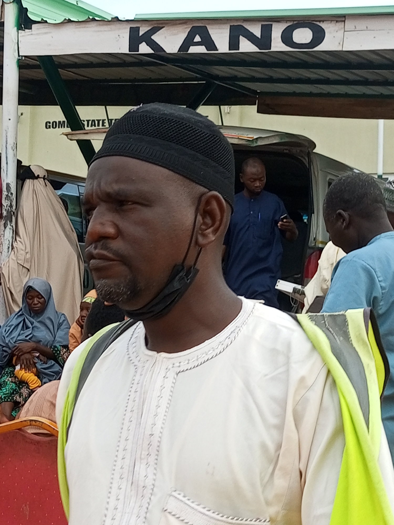 The image shows a group of people, predominantly men, near a building with a sign that reads "KANO". A man in the foreground, wearing a black cap and a white garment with intricate embroidery, is looking to the right. He has a black mask hanging from his ear and is wearing a neon green and grey vest over his shoulder. In the background, other individuals are visible, some near a vehicle. One man in a blue outfit is looking at his phone. The setting appears to be an outdoor area, possibly a transportation hub or a market, with a covered structure overhead. The overall scene suggests a moment of activity and interaction among the people present.