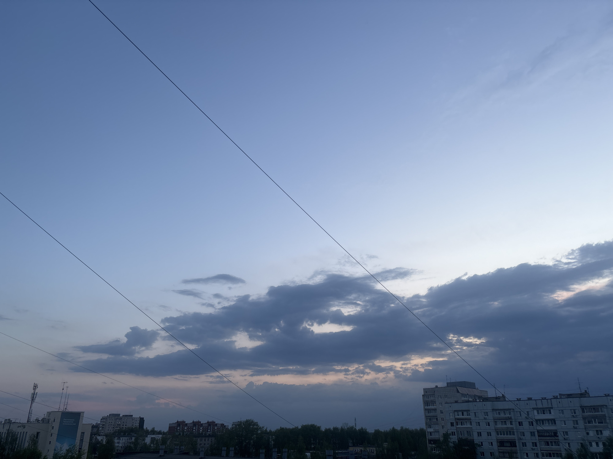 The image presents a late-day cityscape in what is likely Chaikovsky, Russia. The sky is a gradient of blue to greyish-white, with overcast formations dominating the lower portion of the view. Three prominent electrical wires traverse the sky diagonally. Below, urban structures of varying heights are visible, including apartment buildings and other low-rise buildings. Some trees are interspersed among the structures. The buildings appear to be typical residential and commercial structures of the region. In the lower left corner, a sign on a building is partially visible, while rooftops and antennae are seen against the horizon. The overall scene is still and absent of visible human activity.