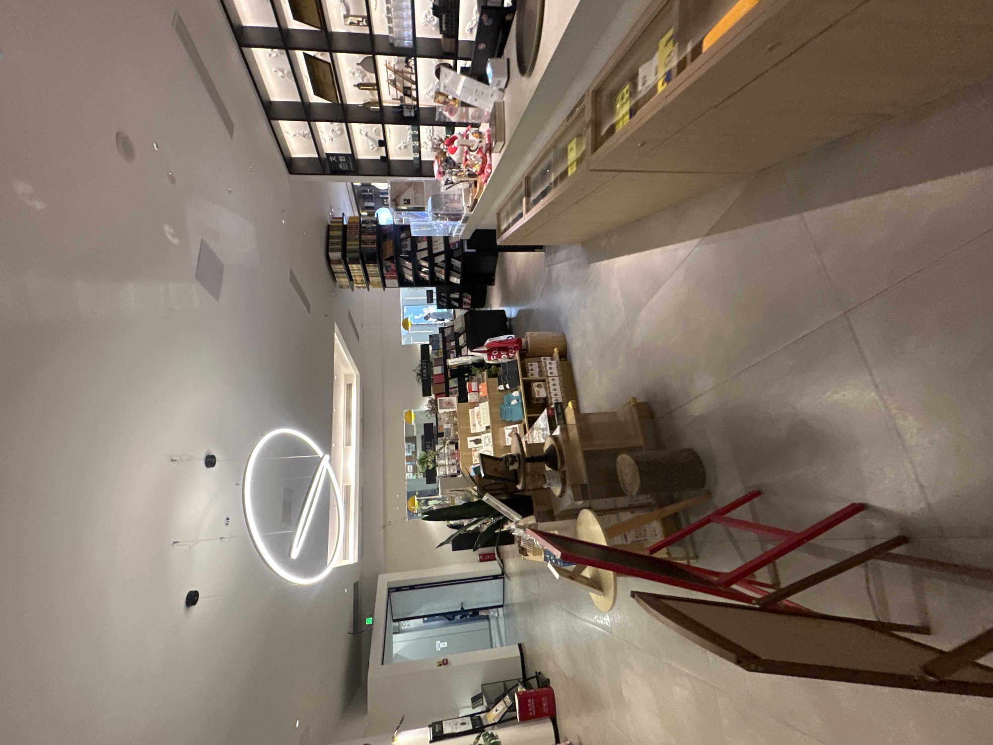 The image depicts the interior of a modern bookstore located in Chongqing High-tech Zone, China. The scene is well-lit with recessed ceiling lights and a prominent circular pendant light fixture. The floor is made of light-colored tiles.
In the foreground, two easel-style signs with wooden frames are positioned on the left, one tilted. Behind these, a section of a wooden display shelf is visible, featuring various books and decorative items, including a small wooden stump.
The midground reveals more of the bookstore's layout. To the left, a doorway or entrance is visible, with a green sign above it. Further into the store, rows of bookshelves are arranged, some filled with books, others displaying art or cultural items. A counter with display cases is situated on the right side of the image, showcasing merchandise. On top of the counter, there are decorative items, including what appear to be figurines.
In the background, more bookshelves and display units extend into the depth of the store. The overall environment suggests a retail space designed for browsing and purchasing books and related merchandise, with a clean and contemporary aesthetic.