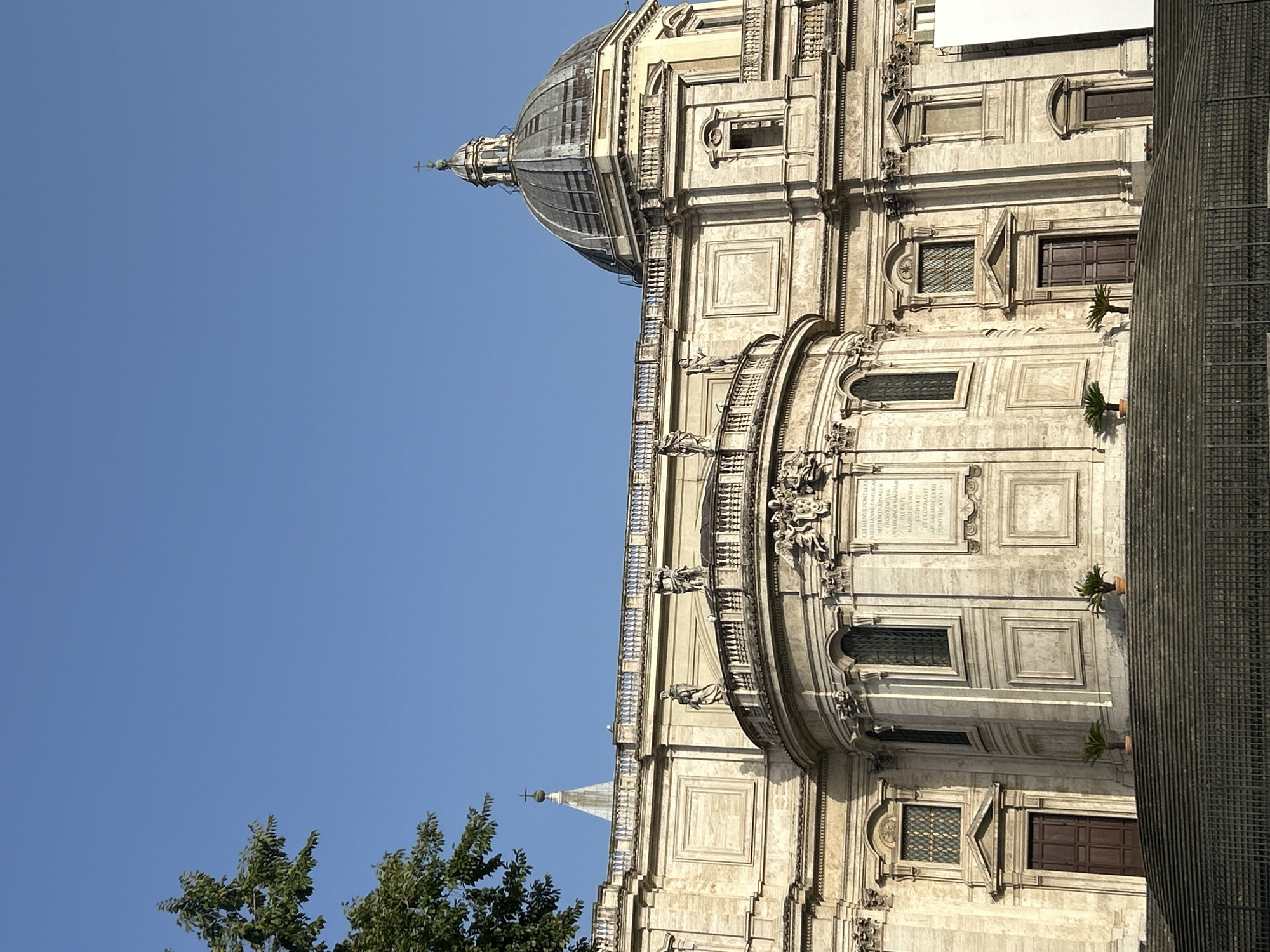 A low-angle, full shot captures the exterior of the Basilica di Santa Maria Maggiore in Rome, Italy, under a clear blue sky. The large, ornate building features classical architecture with detailed stonework, columns, and statues. A prominent dome caps the right side of the structure, and a smaller spire rises from behind the facade on the left. The building's facade is adorned with various windows, doorways, and decorative elements, including a semicircular portico with statues. A wide, stone staircase leads up to the main entrance, with two potted plants on either side of the entrance. A metal fence is visible at the bottom of the stairs. The left side of the image is partially obscured by a lush green tree. The lighting suggests it is daytime, with shadows cast by the architectural elements.
