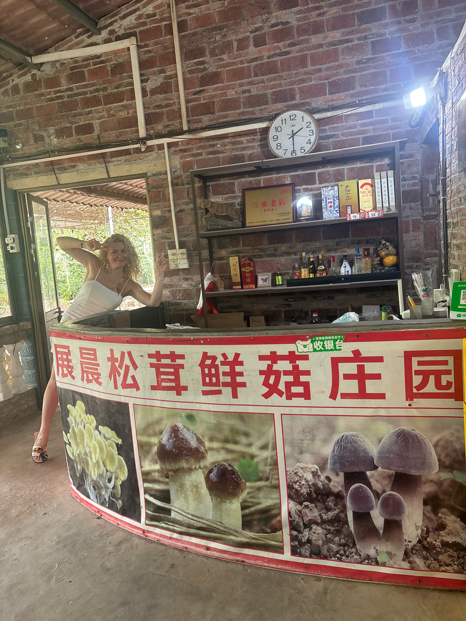 In a rustic, open-air establishment in Sanshui District, China, a woman with blonde, curly hair and wearing a white slip dress and high heels poses provocatively behind a curved counter. She leans forward, making peace signs with both hands. The counter is adorned with a large banner featuring images of various mushrooms and Chinese characters. Behind her, shelves are stocked with bottles of alcohol and other items, and a clock hangs on a brick wall. The scene appears to be a retail space selling mushrooms and possibly local produce or spirits.