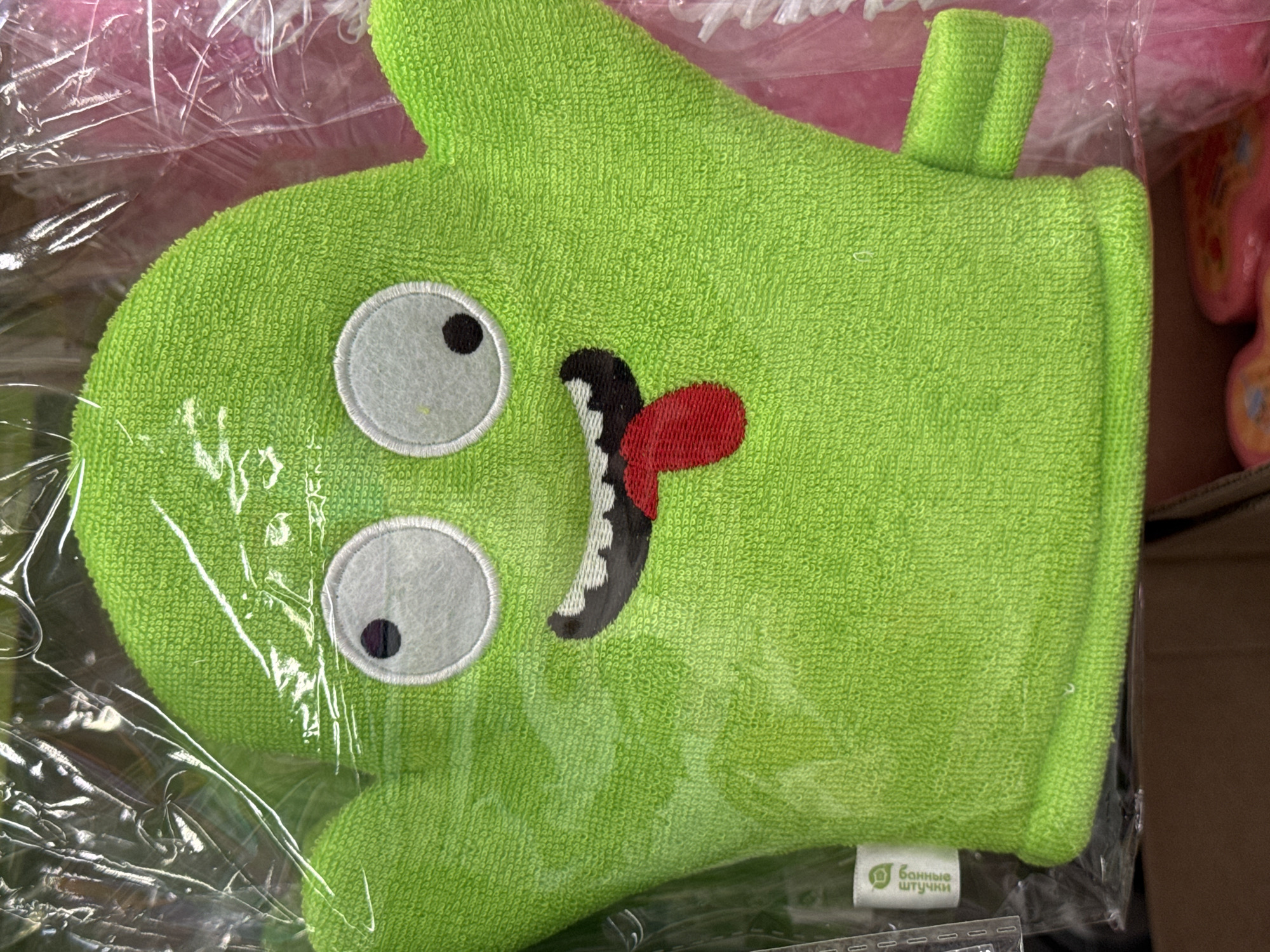 The image shows a close-up of a bright green, terrycloth bath mitt shaped like a cartoon monster. The mitt has two large, white circular eyes with black pupils. Its mouth is open, revealing jagged, black and white teeth and a red tongue sticking out. A small green loop or tab extends from the top of the mitt, likely for hanging. The mitt is enclosed in clear plastic packaging, which has some creases and reflections.
In the bottom right corner of the mitt, a small white tag is visible with green text. The text is in Cyrillic script and reads "Банные штучки" which translates to "Bath little things" or "Bath trinkets".
The background is blurred, but hints of other items are visible. To the left, a pink, possibly fabric or plastic item with a ruffled edge is partially seen, also covered in plastic. To the right, the edge of a cardboard box is visible, and behind it, a blurry pink object with a yellow and black pattern, possibly a toy or another product, can be discerned. The overall lighting is bright, suggesting an indoor setting, possibly a retail environment or storage area. No people are visible in the image. The mood conveyed by the bath mitt's design is playful and whimsical.