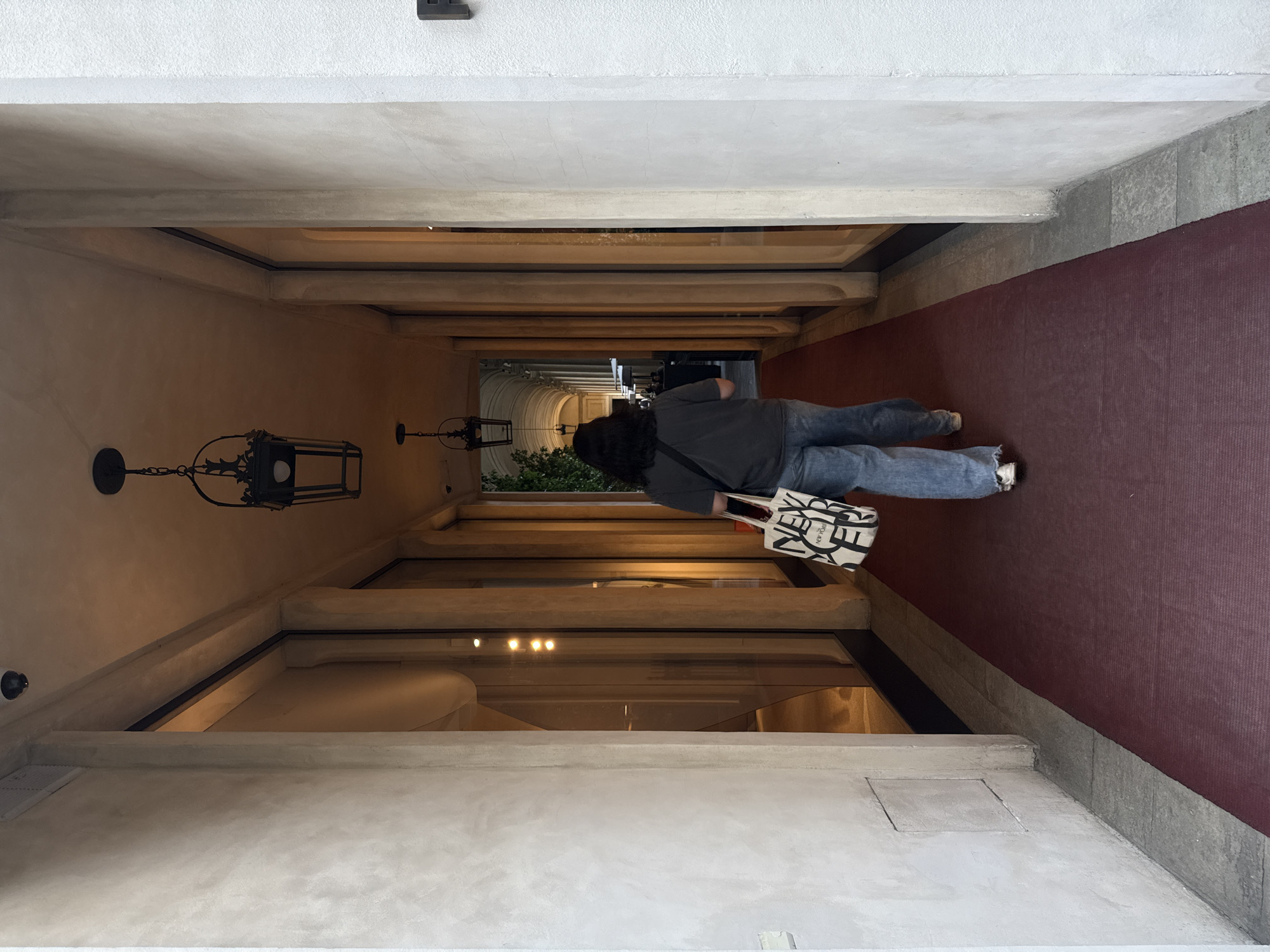 The image shows a person walking down a long, narrow corridor. The person is wearing jeans and a dark-colored shirt, carrying a black and white tote bag. The corridor has a series of archways leading to an open area with green foliage. The corridor is lit by overhead lights, and there are wall-mounted lamps. A red carpet runs along one side of the corridor. The architecture suggests a location in Milan, Italy. The corridor is defined by its long perspective and the repeated archways, creating a sense of depth. The environment is enclosed but leads to an outdoor area visible in the distance.