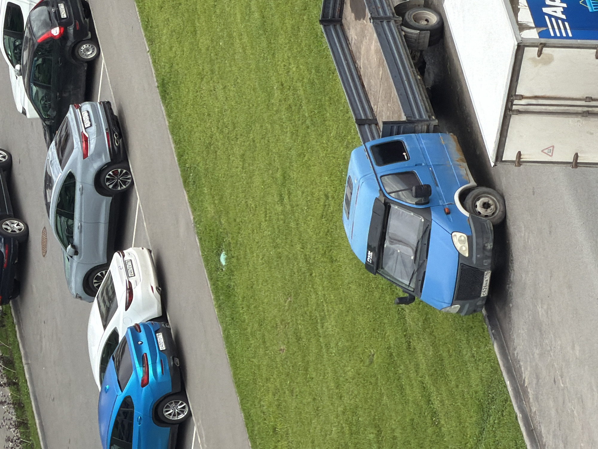 A high-angle outdoor medium shot shows a blue truck with a flatbed trailer parked on a paved surface next to a grassy area. The truck is positioned to the left of the frame and is facing forward. The trailer extends to the right, empty. In the background, to the upper left of the frame, several cars are parked on a paved surface. The cars include a blue coupe, a white sedan, a gray SUV, and a black hatchback. The environment appears to be a residential or commercial area. The lighting suggests daytime.