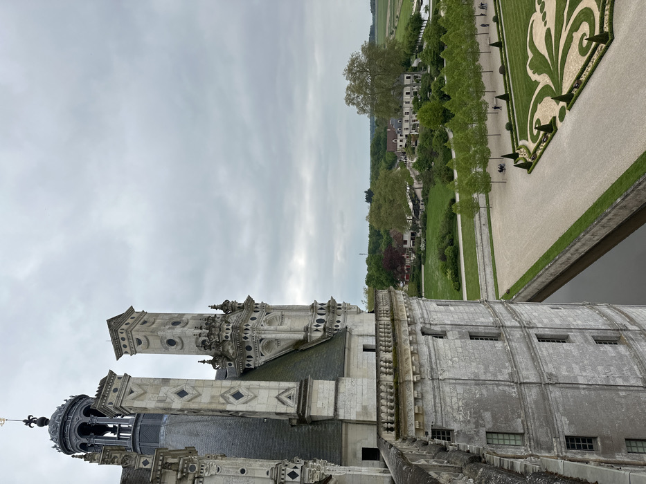 The image depicts a view from the upper level of the Château de Chambord, a Renaissance-era castle in Chambord, France (latitude: 47.616, longitude: 1.518). A portion of the castle's exterior is visible on the left, with a white stone facade adorned with windows, and a curved, ornate parapet. The castle's moat is directly below, with a narrow, rectangular channel of water visible in the foreground. Beyond the moat is a meticulously manicured landscape. A row of trees lines a pathway running parallel to the moat. A large, open space in the foreground is covered in a layer of gravel. This area features a complex design formed by a series of intersecting lines, arranged in a pattern that resembles a fleur-de-lis. There are a few individuals visible in the distance, some are seated on a stone bench while others are standing on the pathway. Beyond the manicured grounds, the countryside spreads out, with a large field to the right and a mix of trees and buildings in the background. The sky is overcast with gray clouds.