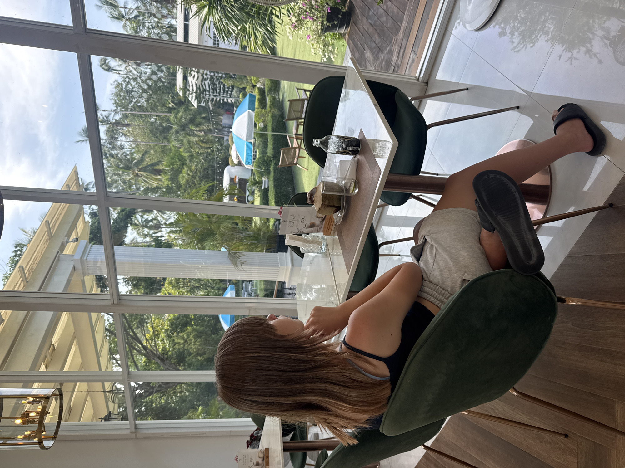 A young woman with shoulder-length brown hair is seated at a marble-top table in a cafe. She is wearing a dark blue tank top and light gray shorts. The cafe has large windows that look out onto a lush green garden with palm trees, two blue and white umbrellas, and some white structures. The woman is looking out the window, with her chin resting on her hand. On the table in front of her, there is a glass, a bottle, a small plate with a slice of cake, and a young coconut with a straw. The cafe interior features modern furniture, including dark green velvet chairs and marble tables. The flooring is a mix of polished wood and white tile.