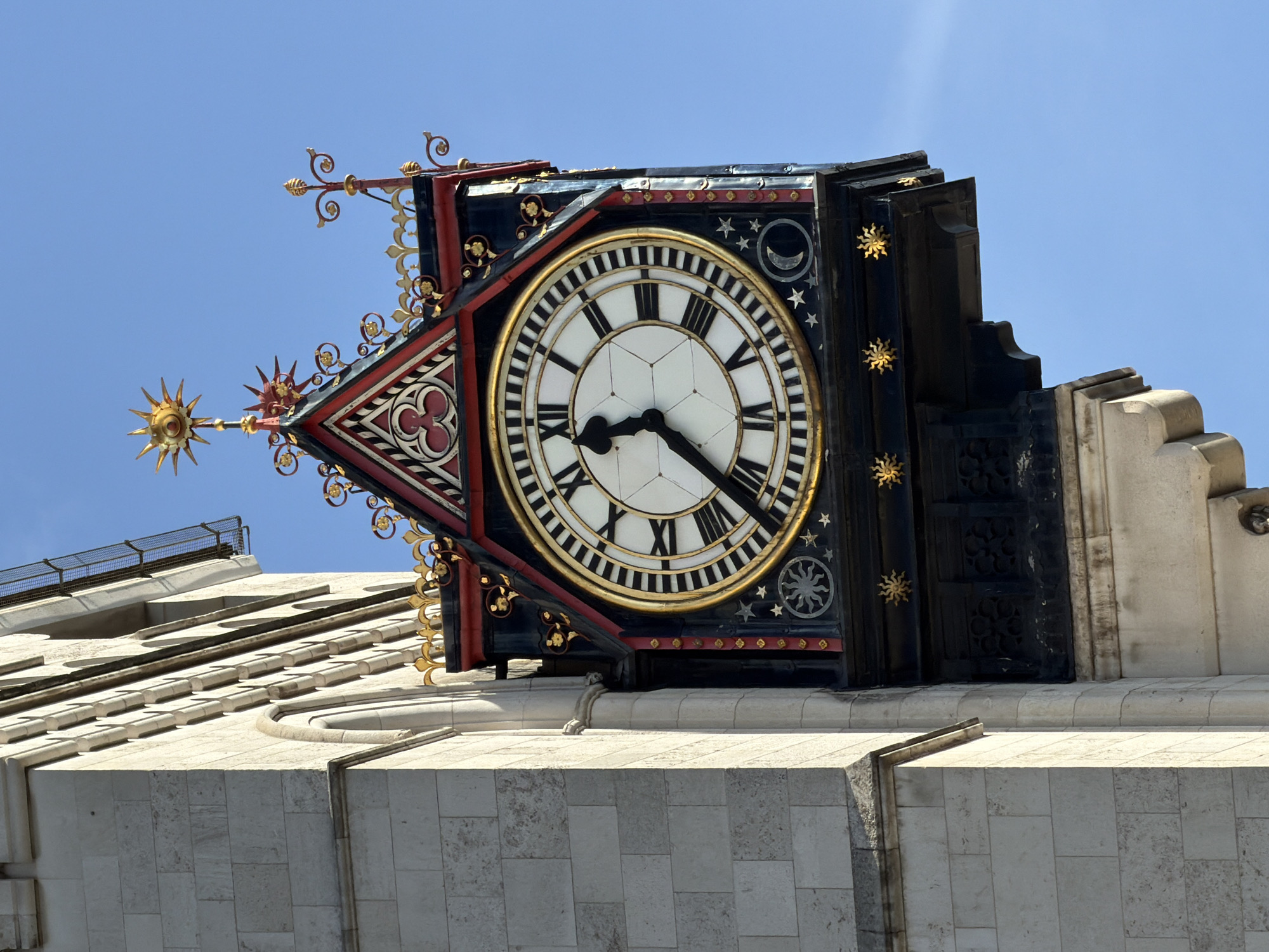 The image shows a close-up of the clock face of the Elizabeth Tower, commonly known as Big Ben, in London, United Kingdom. The clock face is round with Roman numerals and black hands showing approximately 8:20. The clock face is surrounded by intricate gold and red decorative elements. The sky is clear and blue. The tower itself is made of light-colored stone.