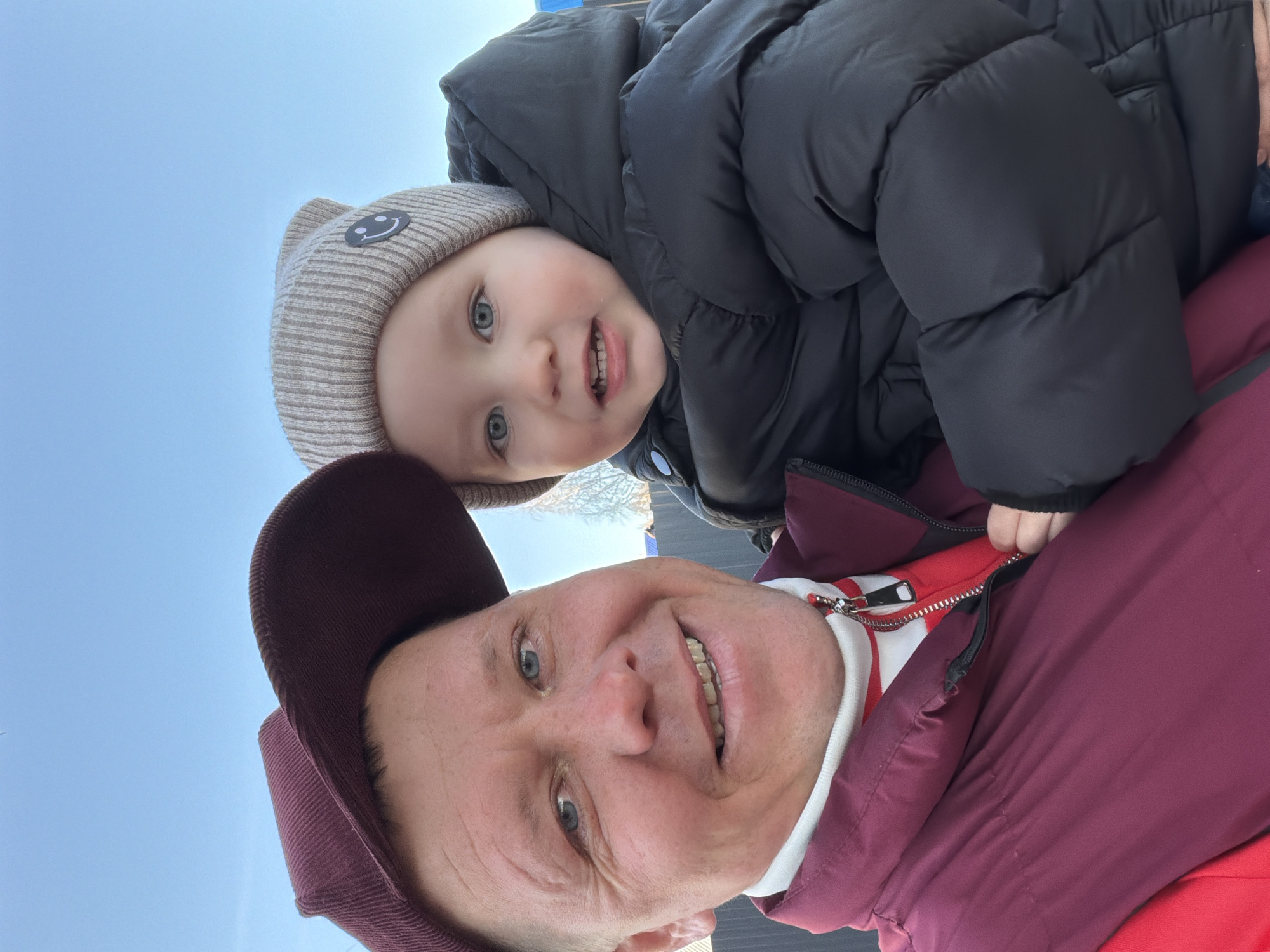 An older man with fair skin is seen from a low-angle perspective, wearing a maroon corduroy baseball cap and a maroon jacket over a white-and-red striped shirt. He is smiling towards the camera. Perched on his shoulders is a small child with fair skin, wearing a grey beanie with a smiley face patch and a puffy black jacket. The child is also smiling. The background is a clear, pale blue sky. The location is likely Chaikovsky, Russia.