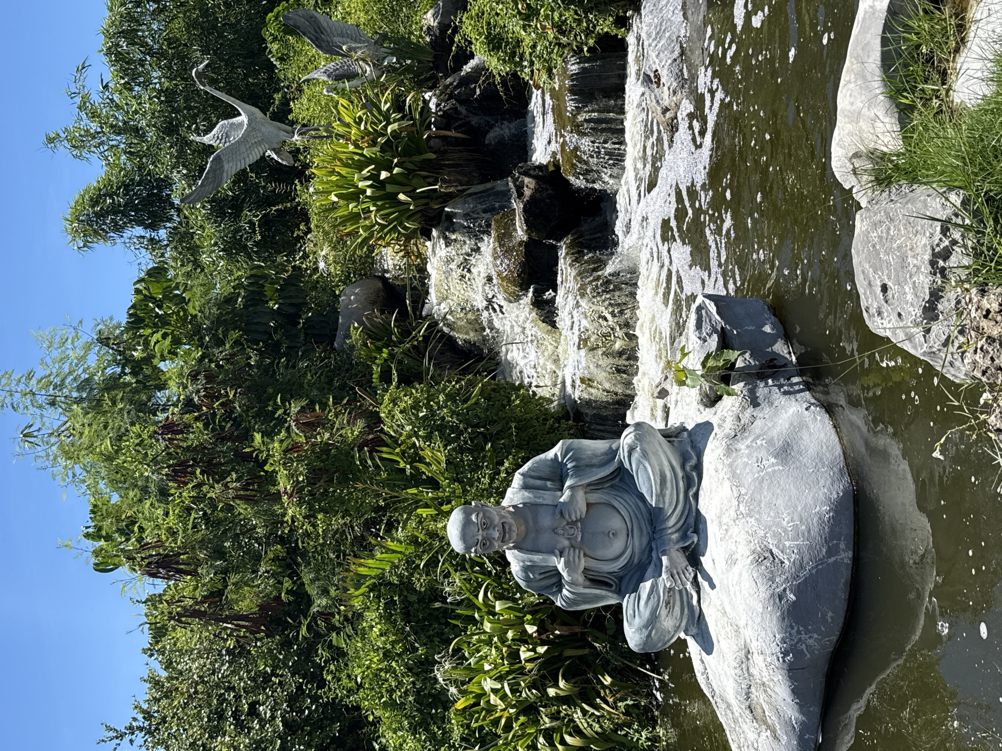 A statue of a smiling, bald, seated man in traditional garb rests on a large stone beside a pond and a waterfall in Bang Pu Subdistrict Municipality, Thailand. Two sculptures of cranes stand among lush green foliage and other natural elements. The sky is clear and blue.