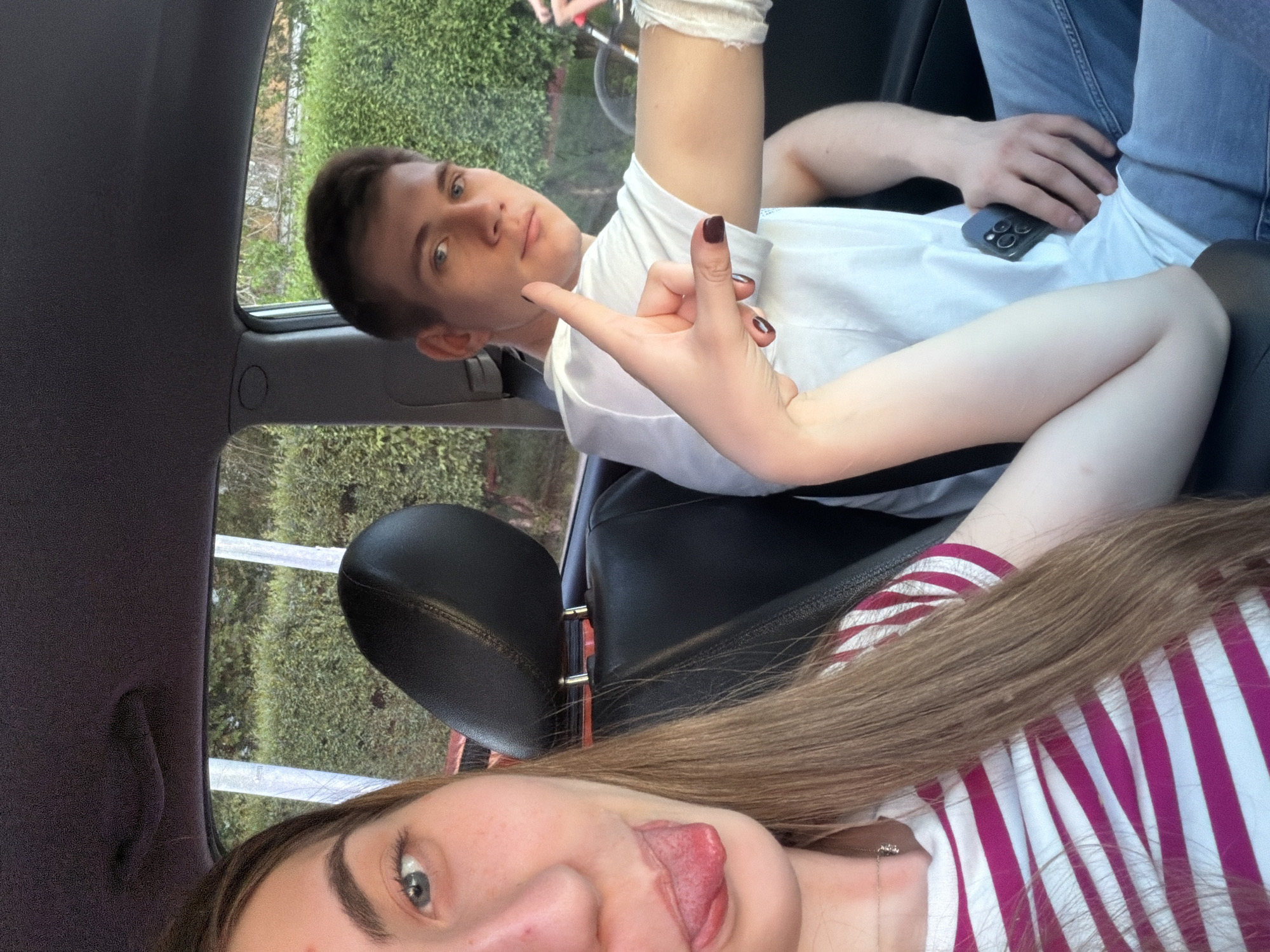 The image captures two young individuals inside a car during daytime, with the composition rotated 90 degrees clockwise. The scene is set in what appears to be Krasnoyarsk, Russia.
In the foreground, a young woman, likely the photographer, is visible from her nose downwards. She has long, light brown hair flowing over her right shoulder and arm, and is wearing a white and pink/red striped striped top. Her tongue is playfully sticking out, and her right hand, with dark nail polish, forms a "rock on" or "shaka" gesture, extending towards the man in the front seat.
Seated in the front passenger seat, a young man looks directly at the camera with a neutral to slightly amused expression. He has short, dark hair and blue eyes, and is dressed in a white t-shirt and blue jeans. A white bandage or wrap is visible on his left forearm, near the wrist. A dark-colored smartphone, possibly an iPhone, rests on his lap.
Through the car windows, lush green foliage, including bushes and trees, is visible, suggesting an outdoor, possibly natural or suburban environment. The overall mood is casual and informal. No discernible text is visible within the image.