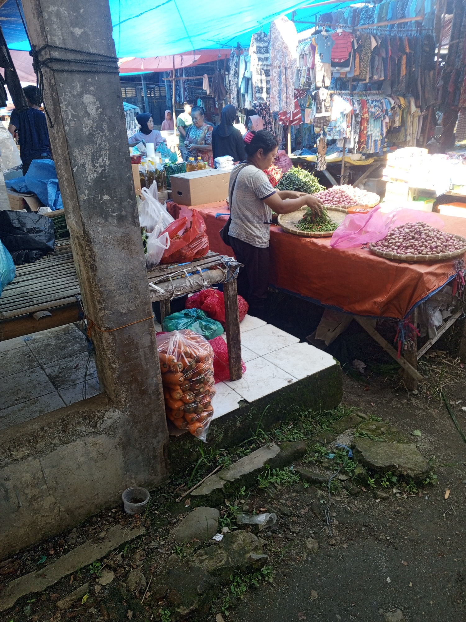 This is an outdoor market scene in Talun Kenas, Indonesia. A woman is sorting produce at a stall covered by an orange tarp, with large baskets of shallots and green chilies on display. Behind her, racks of clothing are visible under a blue tarp. In the foreground, a large plastic bag filled with carrots sits on the ground. Various other items, including plastic bags of produce and boxes, are scattered around the market. The ground is a mix of dirt and pavement, with some vegetation growing in cracks. A concrete pillar is prominent in the left foreground. The atmosphere appears to be that of a busy marketplace.