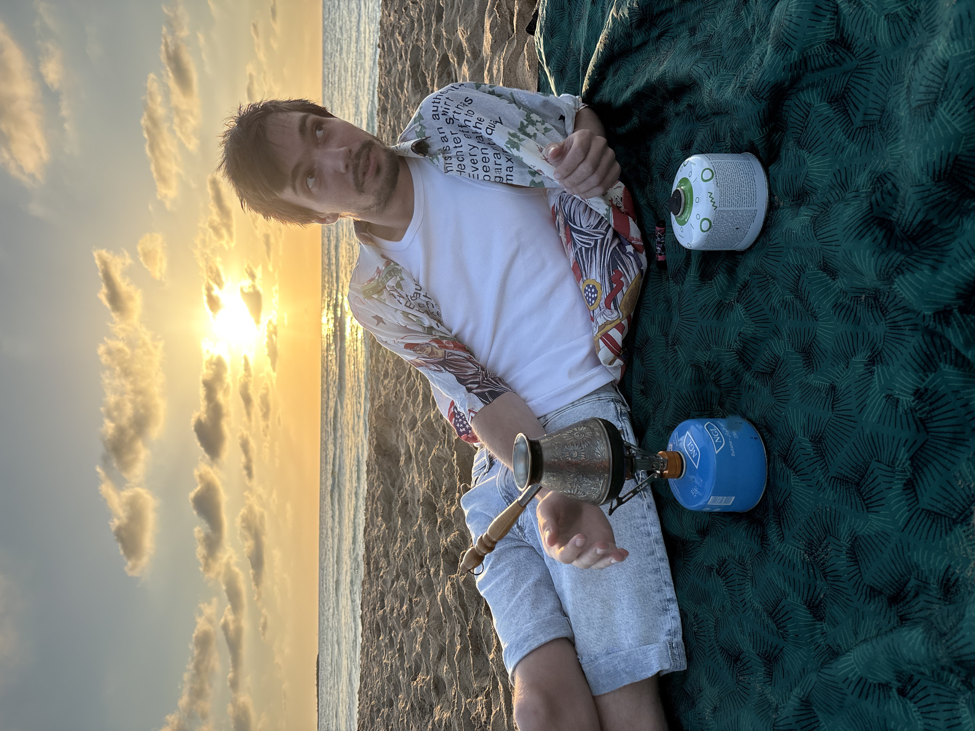 A man sits on a beach blanket at Hof HaCarmel Regional Council, Israel, with the sun setting over the ocean in the background. The man is wearing a white t-shirt, patterned shirt, and denim shorts. He is preparing Turkish coffee on a portable gas stove. There are two gas canisters next to him on the blanket. The sky is filled with clouds reflecting the warm light of the sunset.