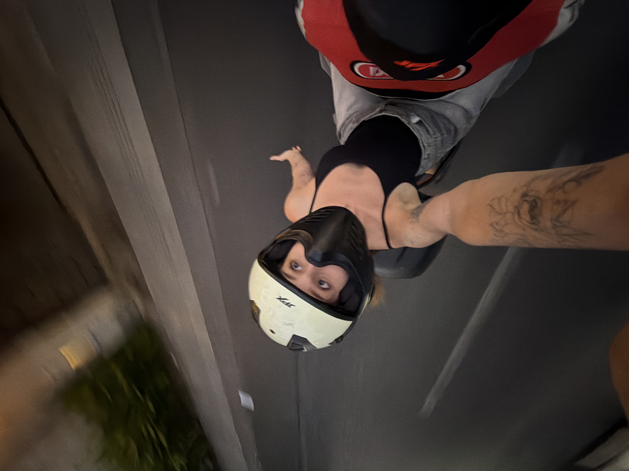 The image depicts a low-angle, first-person perspective of a person riding on a vehicle, likely a motorcycle, in North Kuta, Indonesia. The rider in the foreground, a person wearing a black tank top and grey jeans, is looking upwards towards the camera with a startled expression. They are wearing a cream-colored helmet with black accents and a chin guard. A tattooed arm is extended, holding the camera. The background shows a blurred asphalt road with some foliage and a wall or structure visible on the left side, indicating an urban or suburban environment. The motion blur suggests the vehicle is in motion.