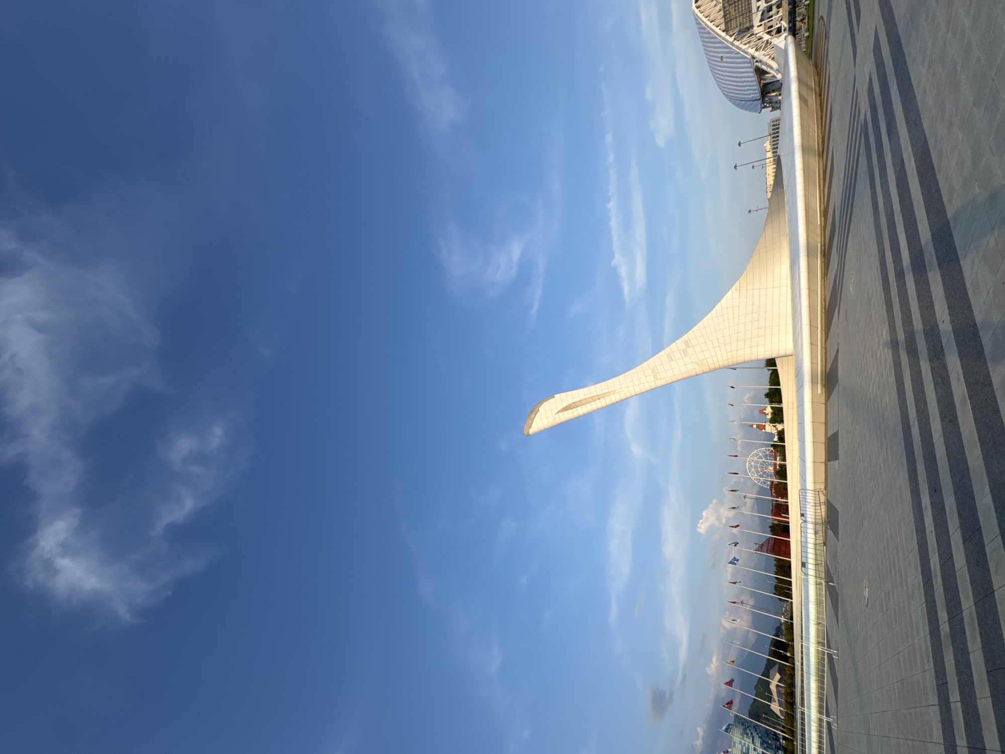 This is a wide-angle, eye-level shot of a modern architectural landmark in Sirius, Russia. The main subject is a large, white, curved structure that resembles a stylized flame or swan, rising towards a clear blue sky with scattered white clouds. The structure has a smooth, polished surface that reflects the sunlight. In the background, a row of national flags on tall poles stretches across the scene, with mountains visible beyond them. Further to the right, a large, white, arched building with intricate structural details can be seen, likely a stadium or arena. The foreground consists of a paved plaza with a geometric pattern of gray and dark gray tiles, casting distinct shadows from an unseen light source. There are no people or animals visible in the image. The overall impression is one of grandeur and openness, with a focus on modern design set against a natural landscape.