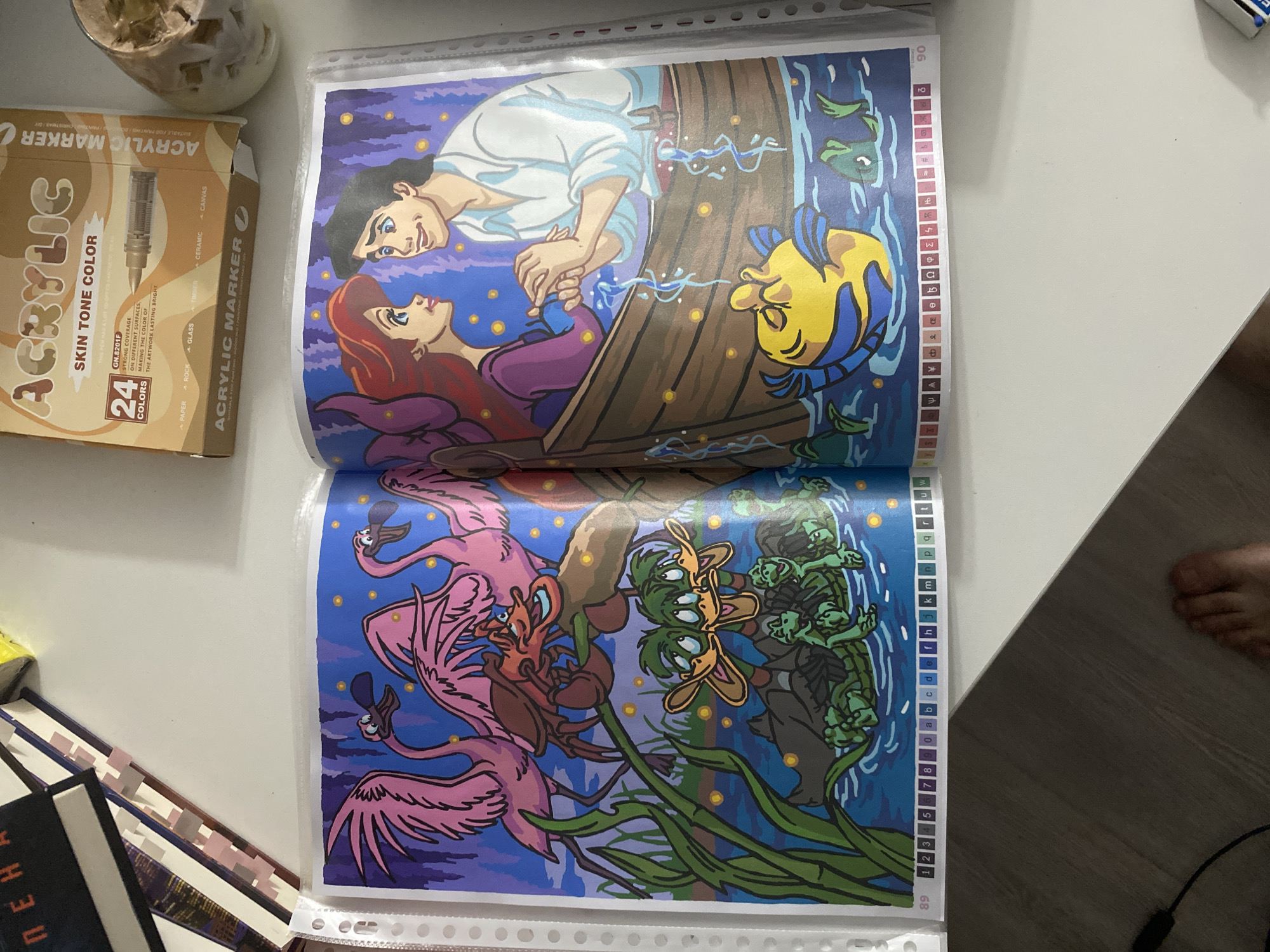 The image shows an open coloring book with illustrations from Disney's "The Little Mermaid." The left page depicts several cartoon flamingos, a crab, and duck-like creatures in a body of water with reeds. The right page features Ariel and Prince Eric in a rowboat, with Flounder swimming below. The coloring book is placed on a white surface, possibly a table, alongside other items. In the upper left corner, partially visible books with spine text are present. To the upper right, a box of "ACRYLIC SKIN TONE COLOR" markers and a decorative cup with ice or a beverage are visible. The lower part of the image shows a white table surface and a dark wooden floor. A human foot is visible on the floor in the lower right corner. The image does not contain any information that would allow for determining its location in the city of Łódź, Poland.
