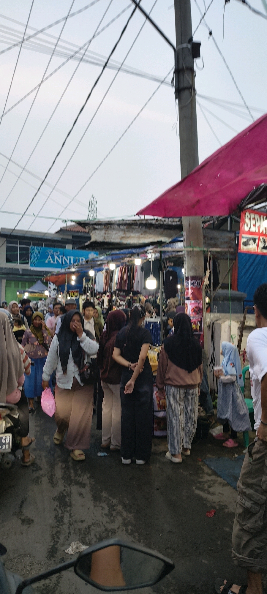 The image depicts a busy street scene at what appears to be a market in Trusmi Wetan, Indonesia. The foreground features the side mirror and handlebars of a motorcycle, suggesting the image was taken from a rider's perspective. The middle ground is filled with a crowd of people, many wearing headscarves, navigating through stalls set up along the road. Clothing items are displayed on racks, and various goods are visible on tables. A prominent red canopy shades a section of the market. In the background, electrical wires crisscross overhead, and buildings line the street. A sign with the word "ANNUR" is visible on one of the structures. The ground is paved and appears wet in places, possibly from recent rain. The overall atmosphere is that of a lively outdoor market.