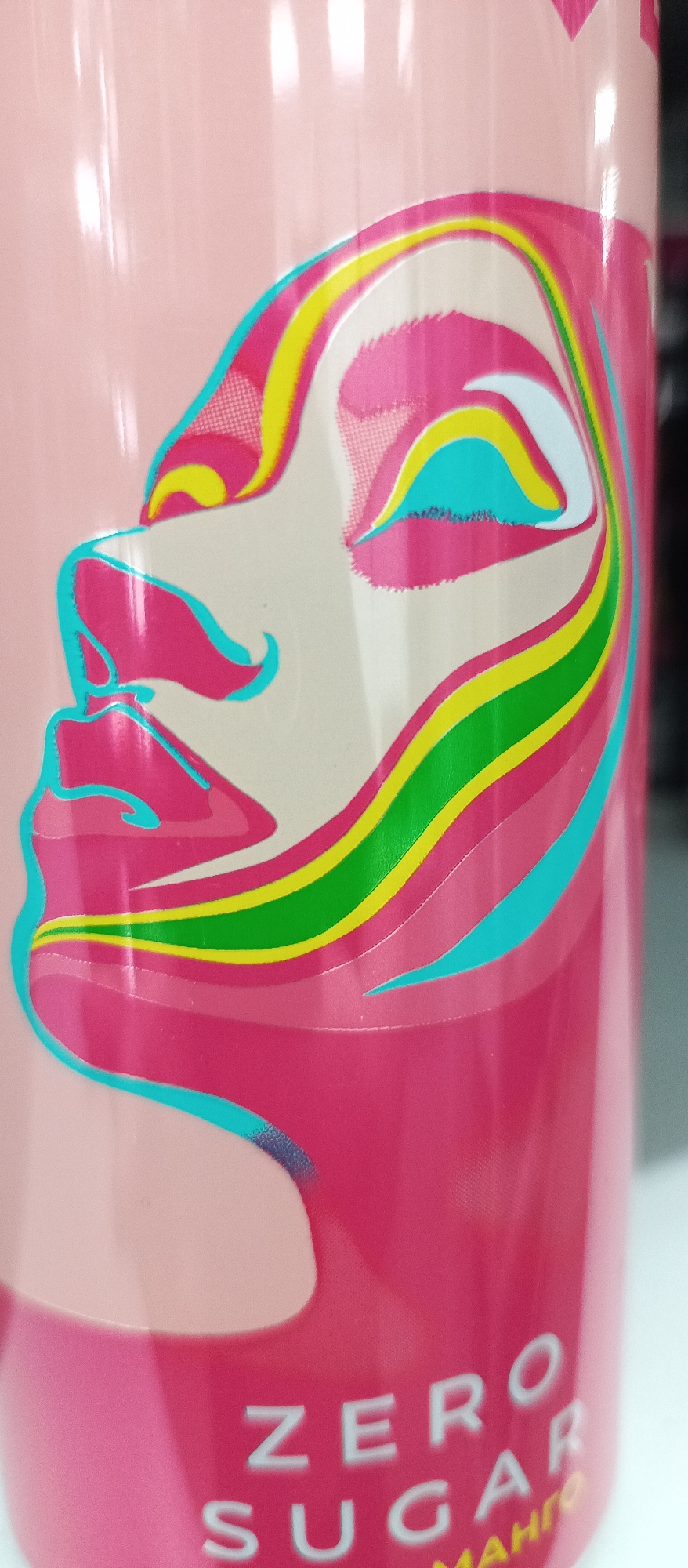 This is a close-up, vertically oriented photograph of a pink can. A colorful, abstract graphic depicting a face in profile is prominently displayed on the can. The face is rendered with stylized, wavy lines in shades of pink, yellow, turquoise, and green, with areas of pale skin tone. The background of the can is a glossy pink. Towards the bottom of the can, the text "ZERO SUGAR" is visible in white, bold, sans-serif lettering. Below "ZERO SUGAR," the word "MANGO" is partially visible in yellow. The lighting suggests an indoor setting, possibly a retail environment or a shelf. The location is Unknown, Unknown.