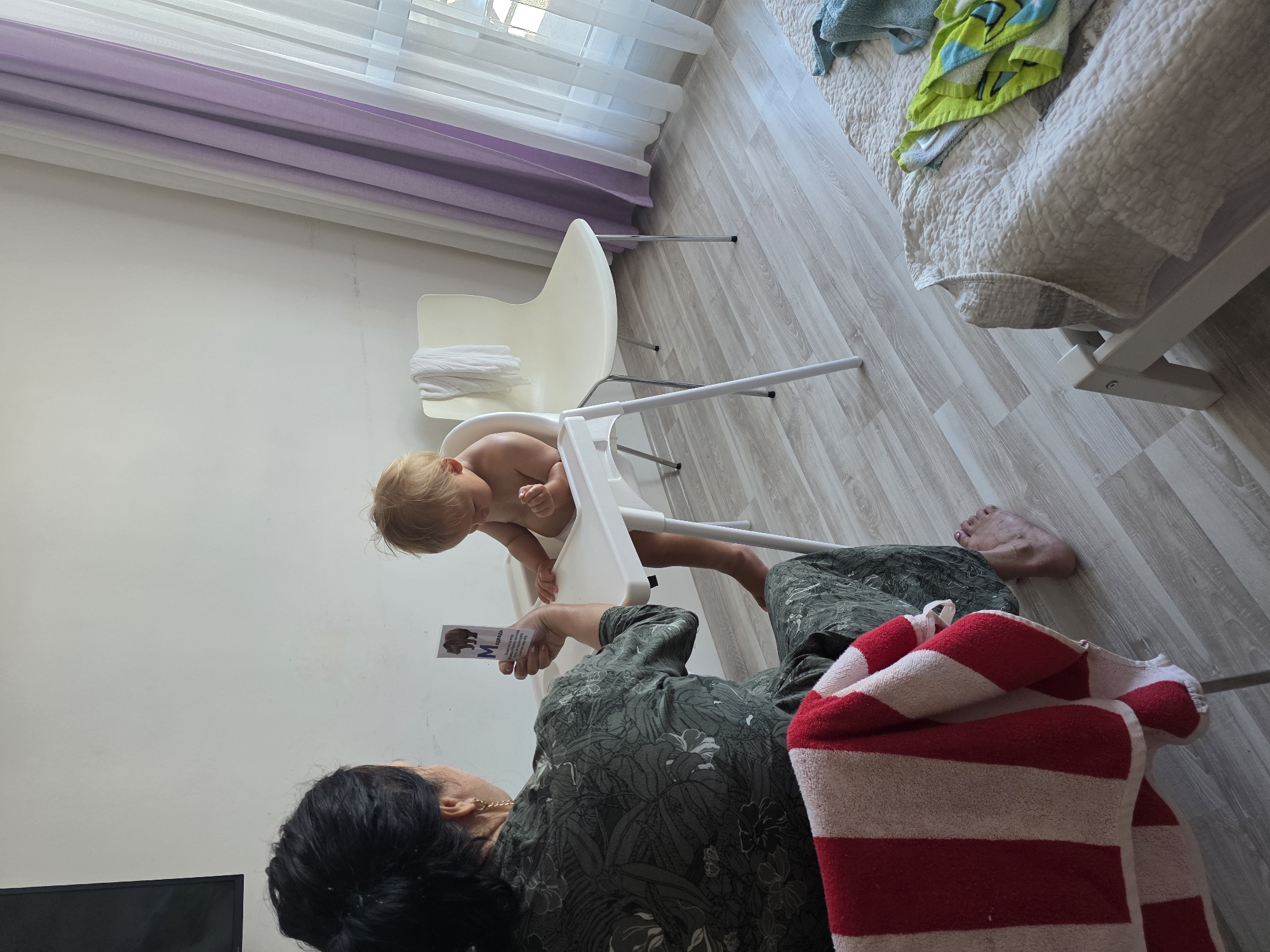 The image depicts an indoor scene at what appears to be a residence in Sochi, Russia. The primary subjects are a baby in a high chair and an adult woman. The baby, with blond hair, is seated in a white high chair. The adult woman, with dark hair and wearing a dark green patterned outfit, is positioned in front of the baby. She is holding up a card with an image of a bear and text, likely a learning aid. A red and white striped towel is draped over the woman's legs and also partially over a leg of the high chair.
The setting includes a light-colored wooden floor and a white wall. In the background, there are sheer white and purple curtains partially covering a window. A bed with a textured light-colored comforter is visible to the right, with a folded green and blue towel on top. The overall lighting suggests it is daytime. The scene appears calm and domestic, possibly indicating a moment of learning or play between the adult and the child. There is a dark rectangular object in the lower left corner, possibly a television screen. The text on the card held by the woman is partially visible and appears to be in Cyrillic script, with the letter "M" prominently displayed.