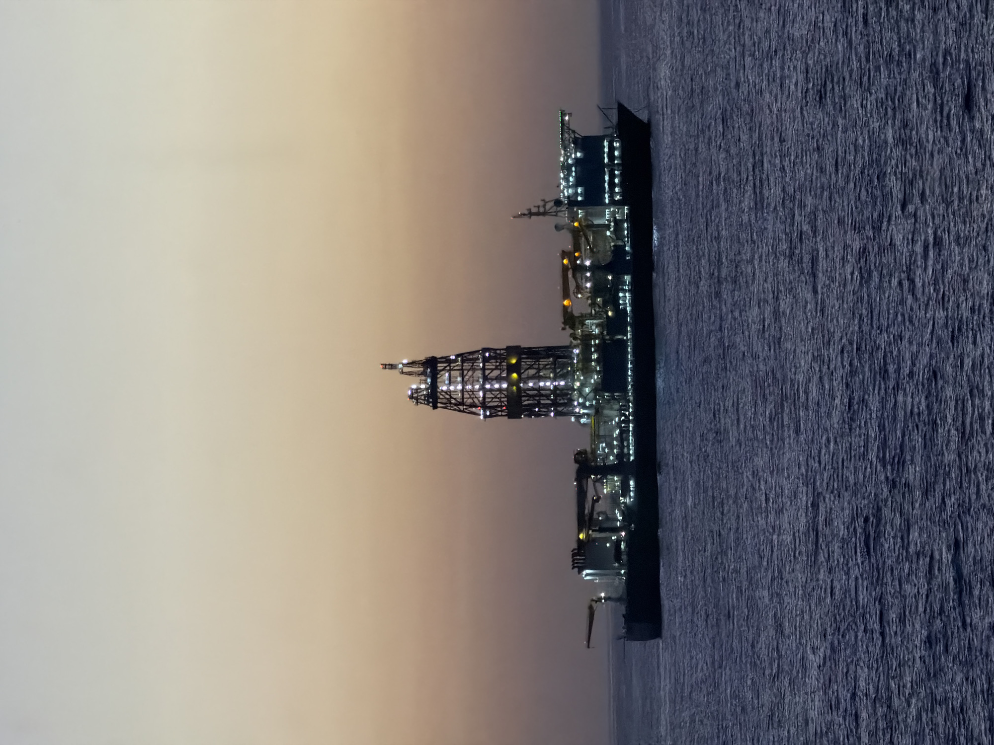 The image displays a large marine vessel, likely an offshore drilling rig or a specialized construction ship, operating at sea. The photograph appears to be taken during dusk or night, as indicated by the dark water and sky, and the numerous artificial lights illuminating the vessel's structure.
The vessel is prominently lit, showcasing a tall, lattice-like tower, possibly a drilling derrick or heavy-lift crane, located midship. Various deck levels, control rooms, and equipment are also visible, brightly illuminated by a multitude of lights. The water beneath the vessel is a dark blue, showing texture on its surface, suggesting either slight choppiness or reflections of the lights.
The sky transitions from a lighter, warm beige or faint orange hue near the horizon, indicative of a sunset or sunrise, to a darker, more muted tone higher up. There are no visible clouds, suggesting clear weather conditions. The scene emphasizes the industrial scale and complexity of offshore operations. No people are visible, and there is no discernible text within the image.