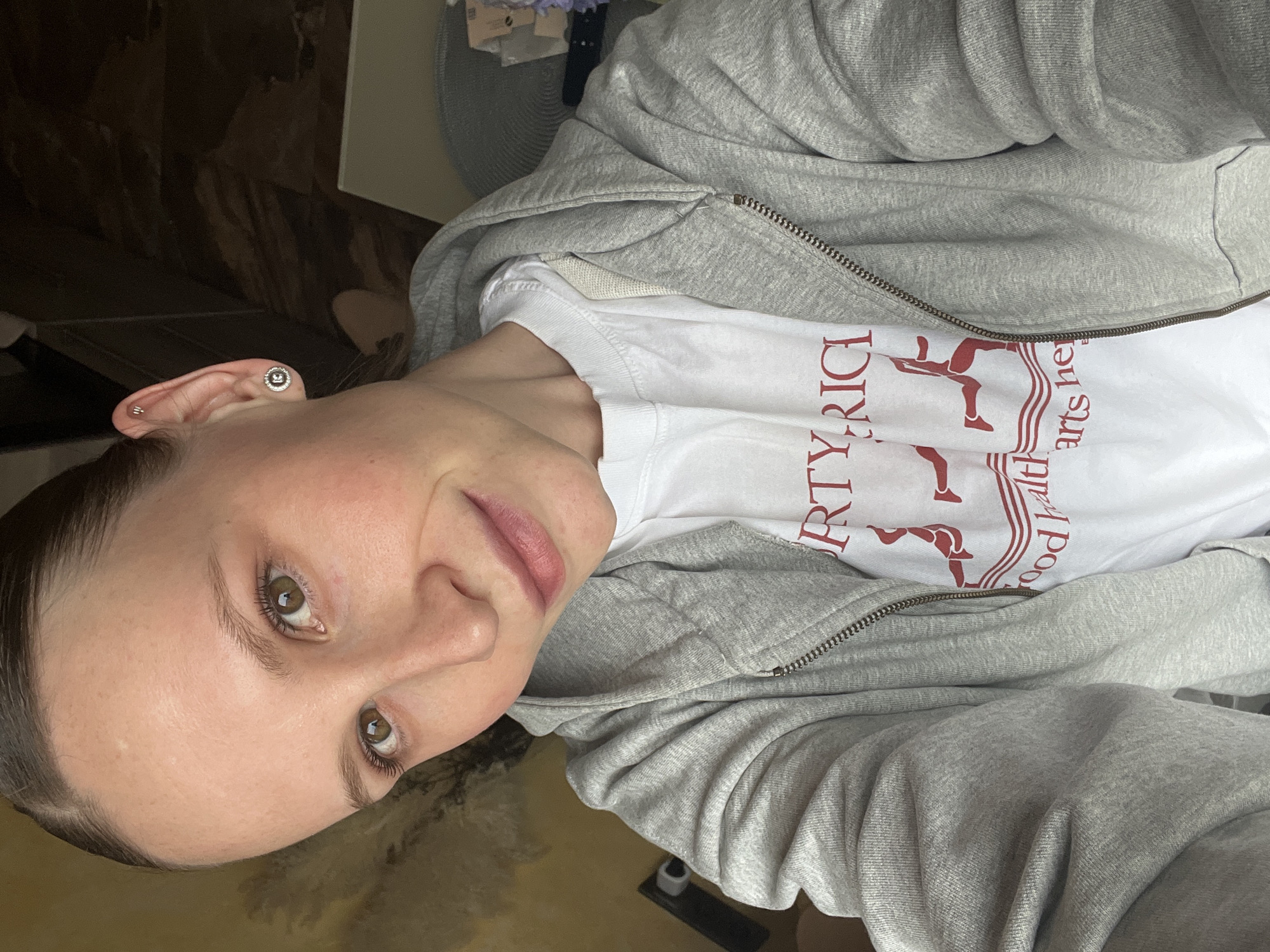 The image features a young adult individual, likely female, captured in a close-up, upward-angled shot. The person is looking directly at the camera with a slight, gentle smile. Their hair is dark and appears to be pulled back, away from the face. They have light skin, brown eyes, and light pink lips.
The individual is wearing a light grey zipped hoodie over a white t-shirt. The texture of the hoodie fabric is visible, showing a soft, possibly cotton or fleece material. The zipper of the hoodie is metallic. On the white t-shirt, there is red lettering and a graphic. Partially visible text includes "RTY RICI" and below it, "ood hearts he" with a stylized graphic of what appears to be a figure in motion, possibly running or dancing.
Notable accessories include a stud earring in each earlobe, one of which is clearly visible and features a dark center surrounded by a metallic, possibly silver-colored, rim with small embellishments. There is also a small, thin metallic bar piercing higher on the ear cartilage.
The background is blurry and indistinct, suggesting an indoor setting. Hints of furniture or walls in dark tones are visible behind the person's head and shoulder. The overall impression is a casual, relaxed pose. The lighting appears to be natural or soft indoor lighting, casting a gentle glow on the subject's face. The context provided indicates the location is Khimki, Russia.