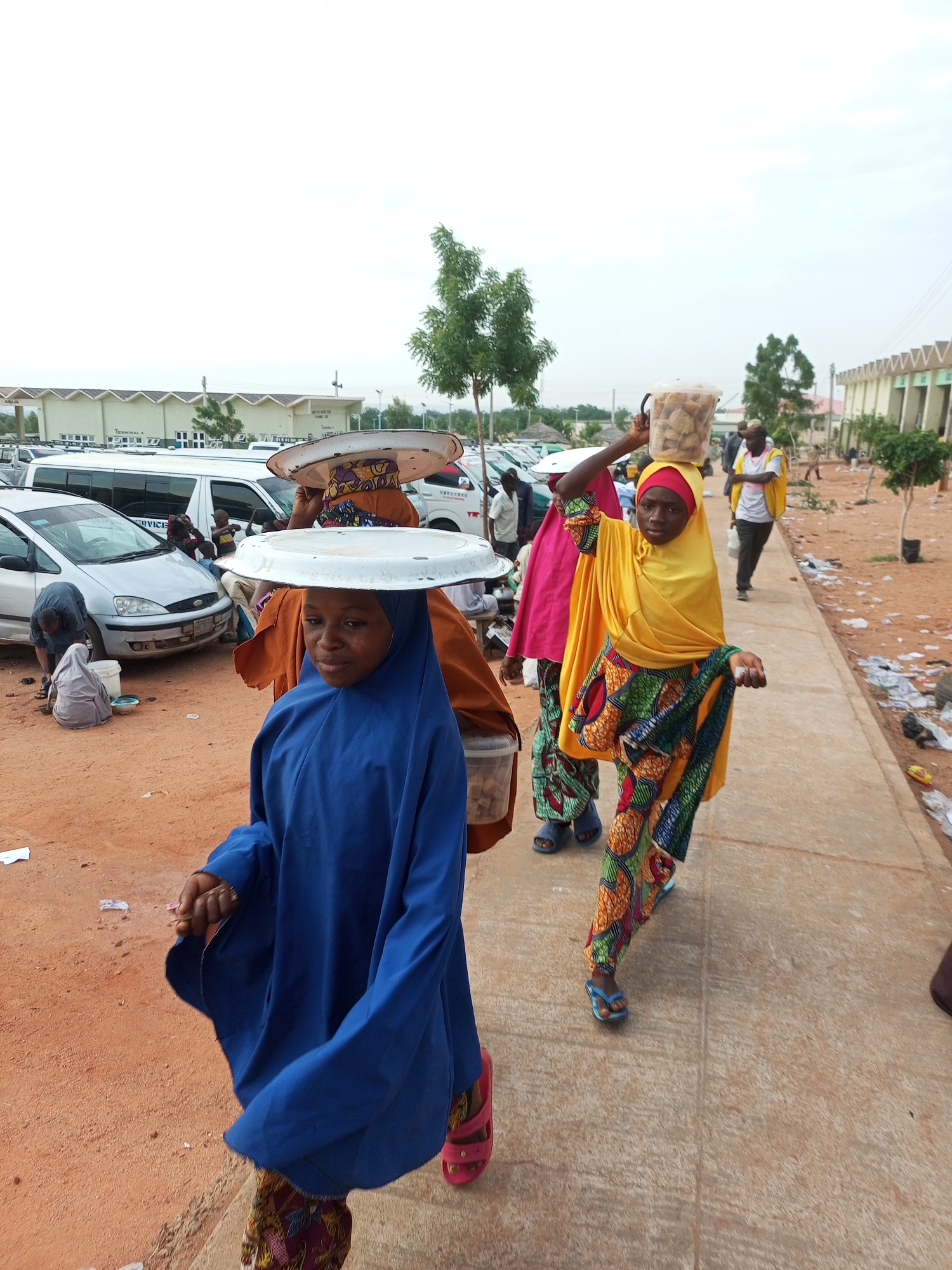 The image depicts a street scene in Gombe, Nigeria, likely near an open-air market or transportation hub. In the foreground, a young girl wearing a vibrant blue abaya and patterned leggings walks towards the viewer, carrying a large, flat metal tray balanced on her head. Her feet are clad in pink sandals. To her right, another young woman, also wearing a headscarf and a brightly colored, patterned outfit, walks away from the viewer, balancing a clear plastic bucket filled with food items on her head. She is wearing blue flip-flops.
Behind them, other individuals are visible. A woman in an orange headscarf and patterned dress is also carrying a large tray on her head. Further in the background, several vehicles, including vans and cars, are parked, suggesting a busy area. The ground is a mix of reddish-brown earth and a paved walkway. Trees are sparsely distributed throughout the scene, and some structures, possibly market stalls or buildings, are visible in the distance. The overall atmosphere appears to be one of daily commerce and movement.