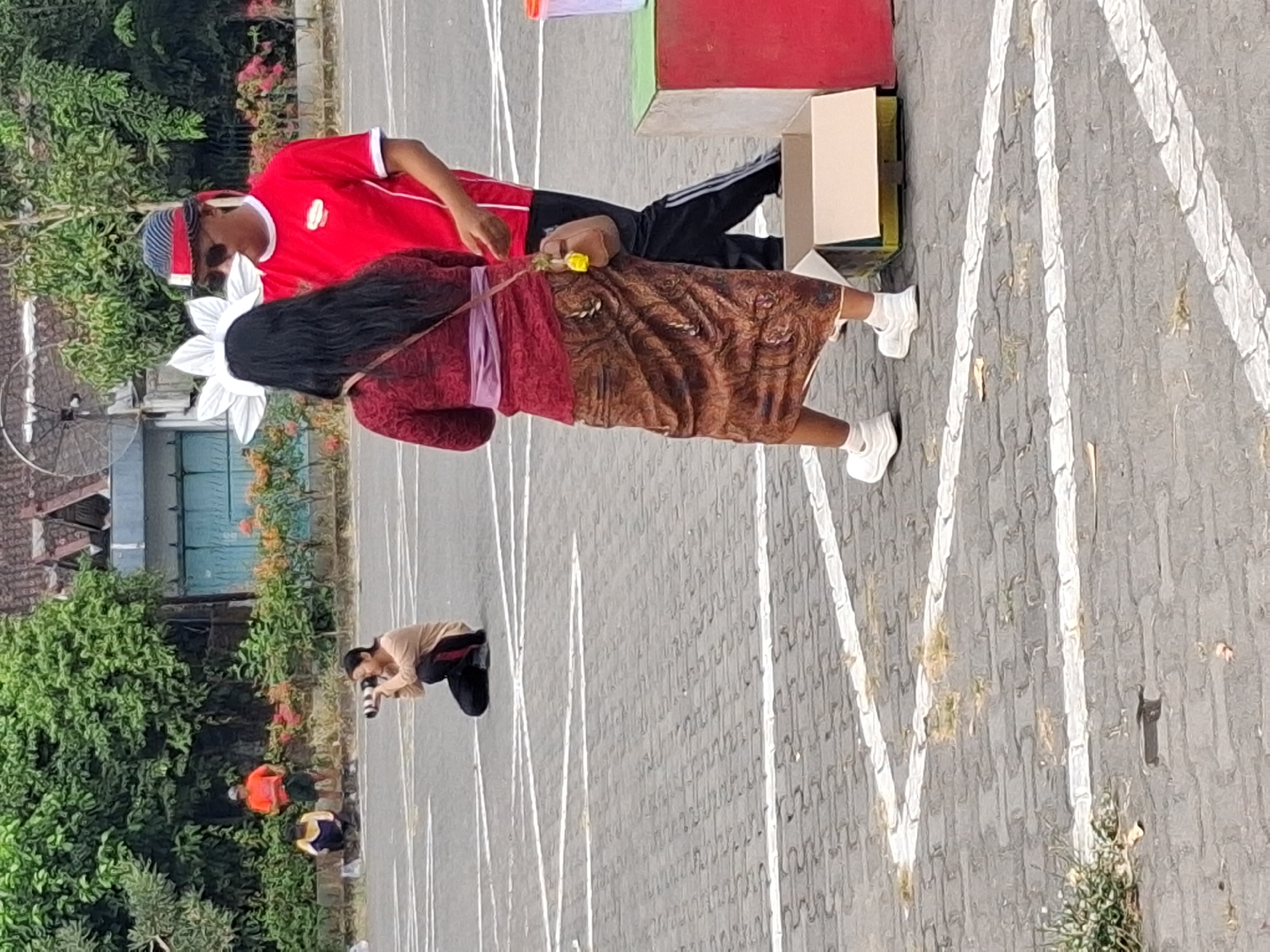 In an outdoor paved area, possibly a parking lot in Surakarta, Indonesia, marked with white lines, a woman in a red long-sleeved shirt and black pants, wearing a red and blue striped headband and sunglasses, is interacting with a woman facing away from the camera. The woman facing away wears a red patterned long-sleeved top with a purple sash around her waist, a brown patterned long skirt, and white sneakers. She has long dark hair and a small brown crossbody bag with a yellow charm. A large white flower-like headdress is visible on her head. In the background, a person is crouched down, holding a professional camera with a large lens, seemingly taking photos. Further back, two individuals are standing near some vegetation and a building. One person is wearing an orange t-shirt and shorts, while the other is wearing a yellow and blue outfit. To the right of the frame, a green and red block structure and an open cardboard box are present. The environment suggests a public outdoor space with some greenery and a building in the distance.