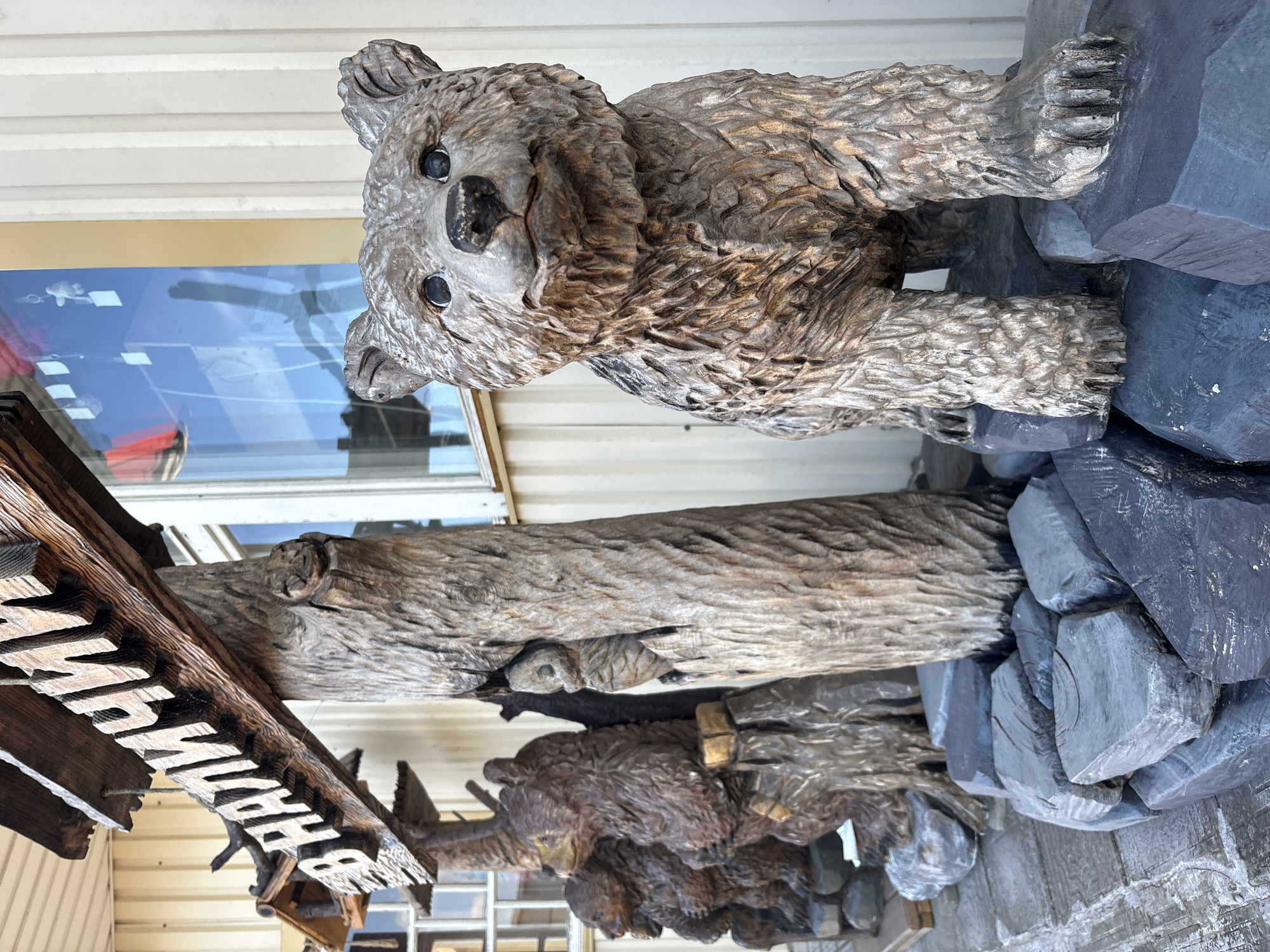 This image captures an outdoor scene in the city of Vyra, Russia, featuring several wooden sculptures of bears and a wooden sign. In the foreground, a detailed carving of a young bear stands on a base of dark gray, sculpted rocks. Behind it, arranged in a line, are more wooden bear sculptures, including what appears to be a mother bear and two cubs, all carved with intricate fur textures and positioned on rough wooden stumps and rocks. To the left, a larger, roughly carved wooden sign with Cyrillic lettering, which reads "В НАЛИЧИИ" (meaning "In Stock"), is suspended above the bear sculptures. The sign is attached to a thick, natural-looking wooden post that is also carved. The background shows a building with cream-colored siding and a window reflecting the sky. The ground is paved with what appears to be grey concrete tiles with a textured surface. The overall impression is one of a rustic, possibly commercial, display of handcrafted wooden art.