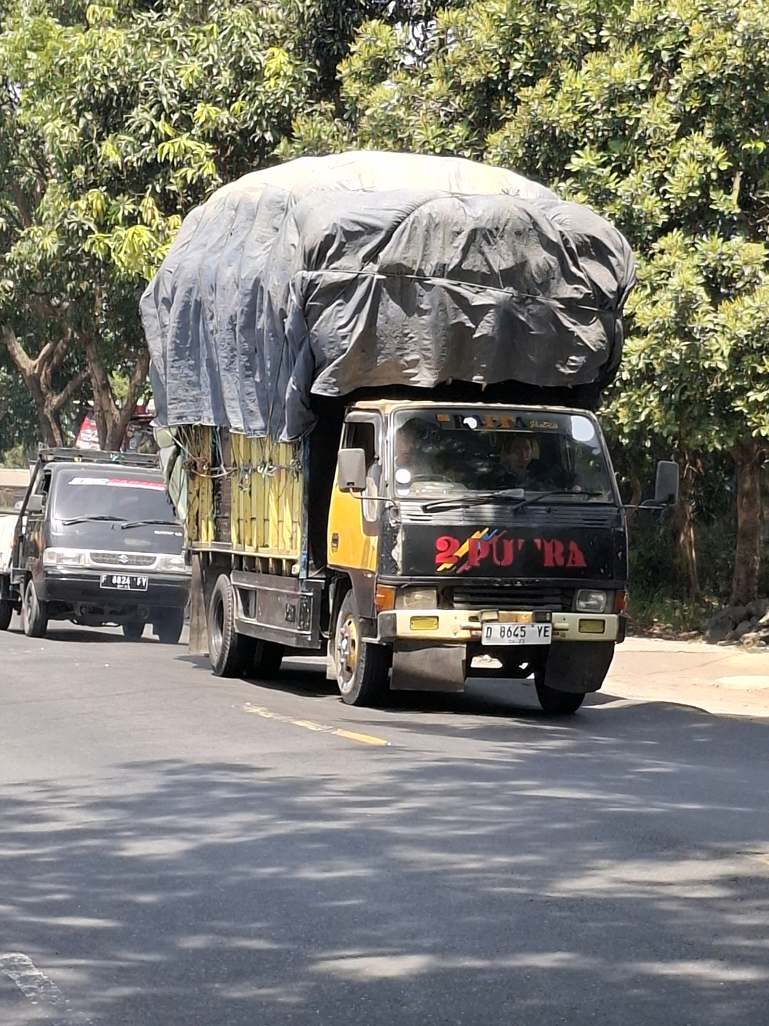 In Cikahuripan, Indonesia, a black Suzuki pickup truck with license plate F 8824 FV is driving on a paved road, partially obscured by a larger yellow and black truck. The larger truck, with license plate D 8645 YE, is carrying an extremely large, bulging load covered by a dark tarp, which is tied down with ropes. The tarp is stretched taut over the contents, creating a rounded, almost balloon-like shape. Two people are visible in the cab of the larger truck. The truck has "2 PUTRA" written in red and yellow on its front. The environment appears to be a semi-urban or rural area with trees lining the roadside. The sun is shining, casting dappled shadows on the road.