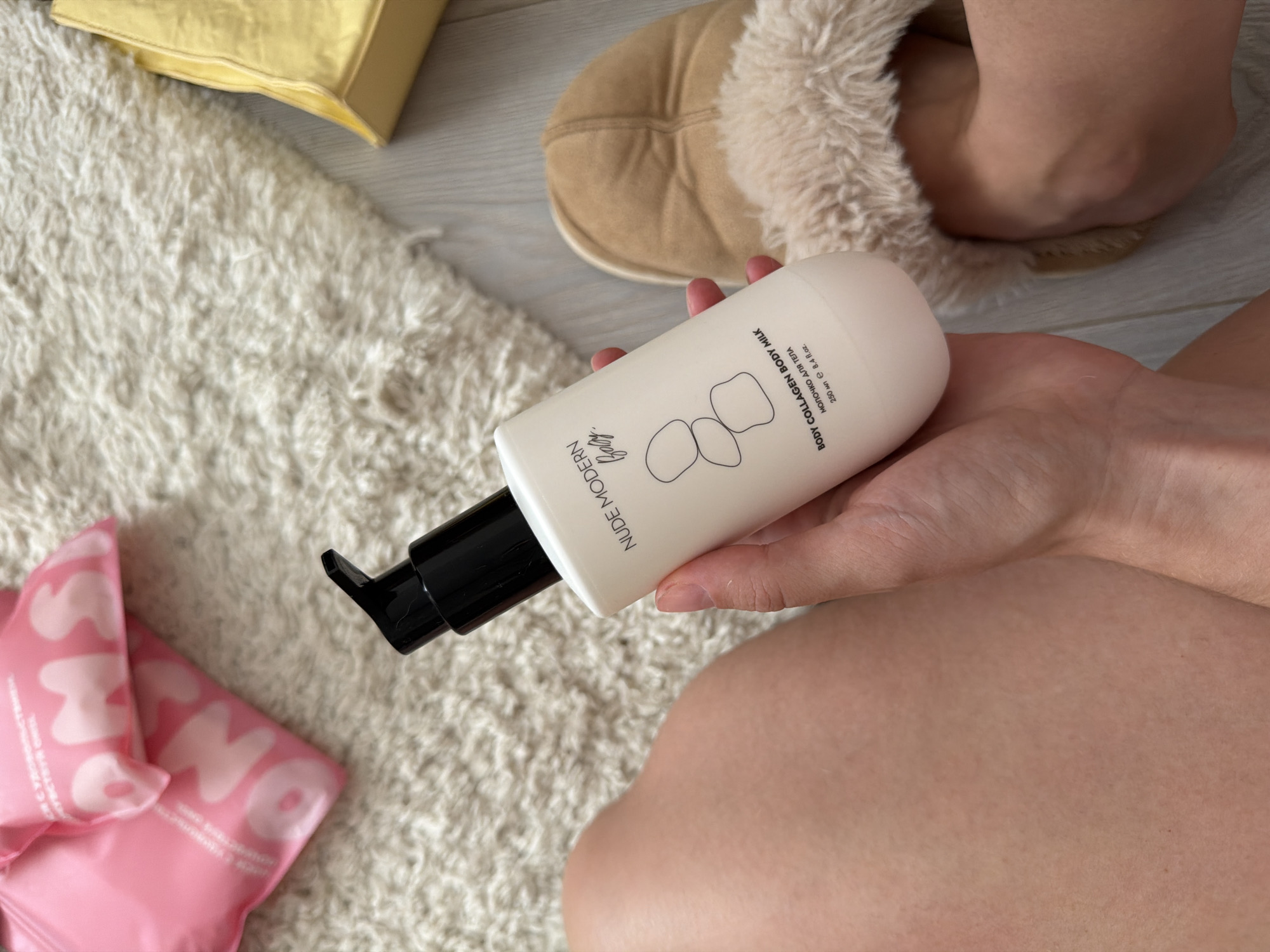 A person's left leg and hand are visible in the foreground. The hand is holding a white bottle with a black pump dispenser. The bottle has text that reads "NUDE MODERN Body Collagen Body Milk" and "МОЛОЧКО ДЛЯ ТЕЛА". Behind the bottle, a fluffy white rug and a yellow bag are visible. In the background, two pink packages with white text are also on the rug. A pair of beige fuzzy slippers with a person's right foot inside them is to the right of the bottle. The scene is indoors. There is no indication of the city of Semiluki, Russia, in the image.