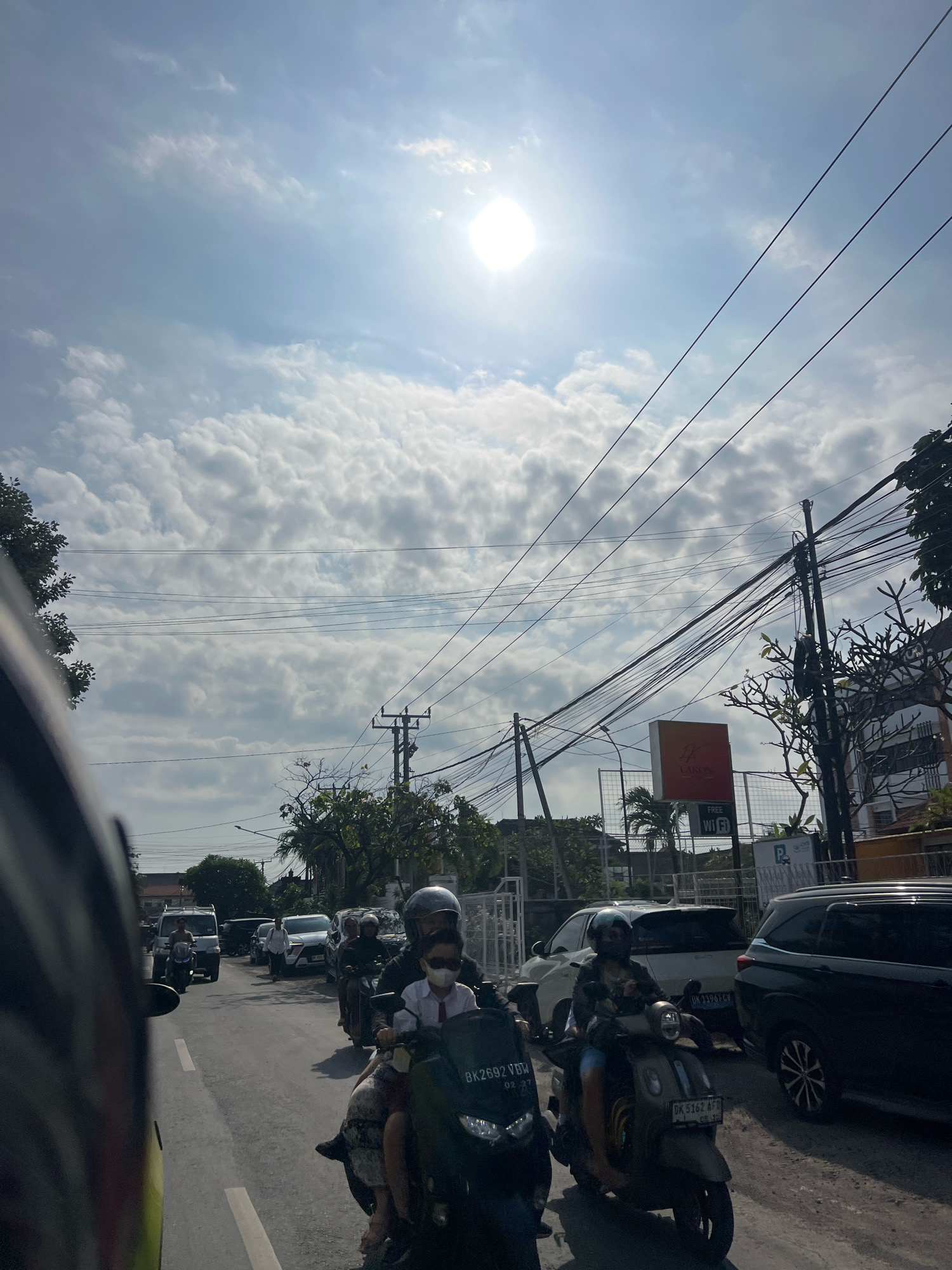 In Denpasar, Indonesia, a busy street scene unfolds under a bright, partly cloudy sky. The sun is a prominent feature, high in the sky and partially obscured by clouds, casting a strong light. The road is congested with various vehicles, primarily motorcycles. In the foreground, a dark-colored car's side and mirror are visible on the left, indicating the photographer is likely in another vehicle.
Several motorcycles are in motion or stopped in traffic. One motorcycle in the mid-ground has two occupants; the rider wears a helmet, and the passenger, a child, has a mask and is wearing a white shirt and a red tie. Another motorcycle further ahead also has two people wearing helmets. Other motorcycles and cars are scattered throughout the scene, indicating moderate traffic.
On the right side of the road, buildings and trees line the street. A tall utility pole with numerous tangled wires crisscrosses the sky, a common sight in urban Indonesian landscapes. A building with a sign that reads "LAKON" and "FREE WI FI" is visible, along with a parking sign and some greenery. The overall environment suggests a bustling urban setting during daylight hours. The road surface is asphalt, marked with white lines.