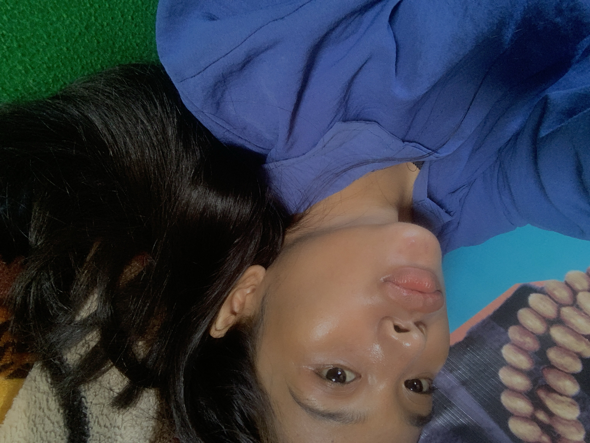 A close-up portrait features a person, likely a young adult female, lying down in an indoor setting in Sungai Meriam, Indonesia. Her head is rotated, presenting her face almost upside down to the viewer, framed by her long, dark hair. She is wearing a blue top or garment. Her expression is neutral, with slightly parted lips and a direct gaze towards the camera. Her skin appears dewy or moisturized.
The background is composed of various textured fabrics: a vibrant green textile is visible to her upper left, more blue fabric matching her top wraps around her upper body, a patterned blanket or cushion with rows of light-colored oval shapes (resembling nuts or beans) is to her right, and a striped brown and cream fabric is beneath her head. The lighting is soft and even, suggesting a relaxed, casual atmosphere, likely during daytime indoors. There is no visible text.