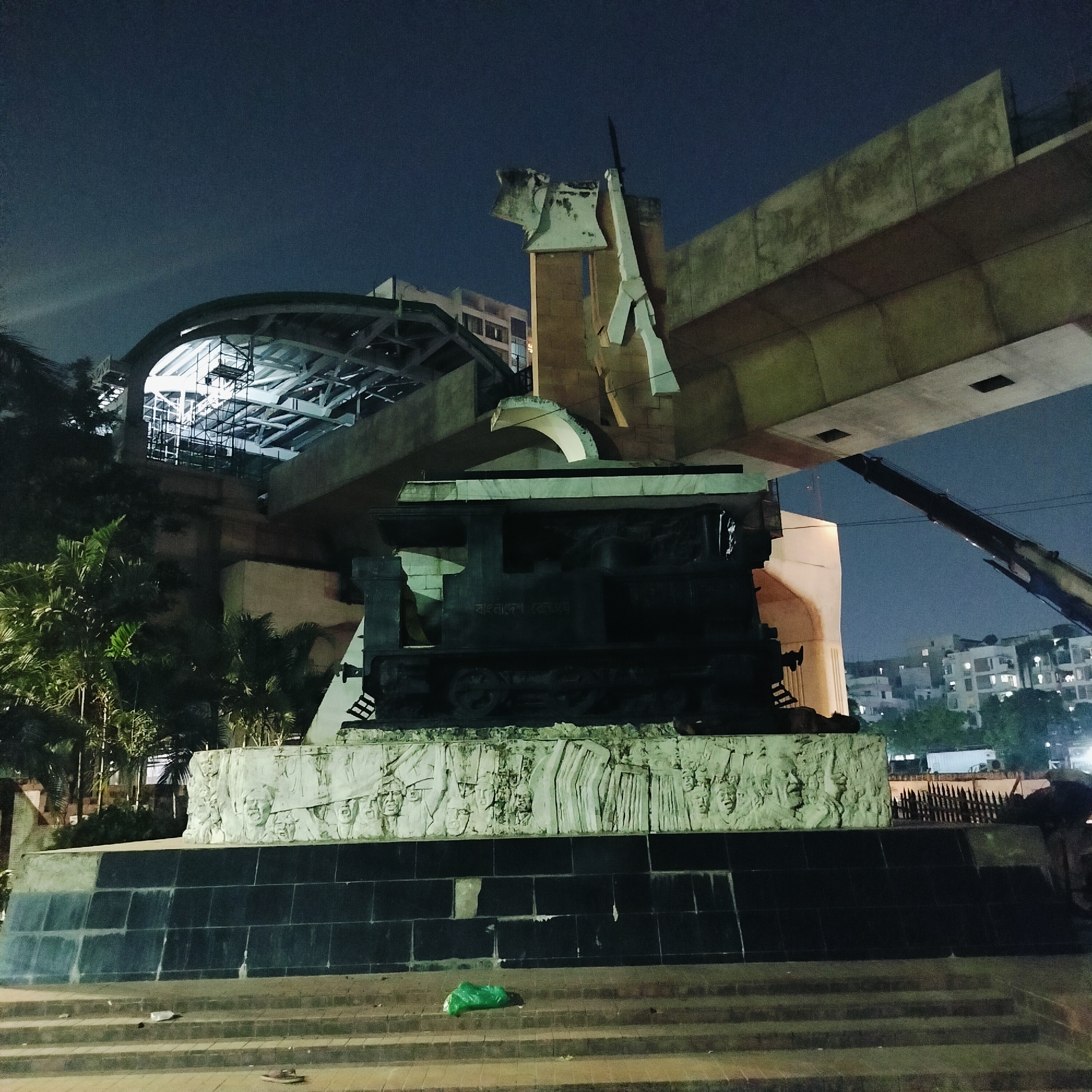 The image shows the Aparajeyo Bangla monument at the University of Dhaka in Bangladesh at night. The sculpture features three figures representing the Bengali freedom fighters. Behind the monument, there is a metro rail structure and other buildings visible in the background. A construction crane is situated on the right side. There is trash on the steps of the monument.
