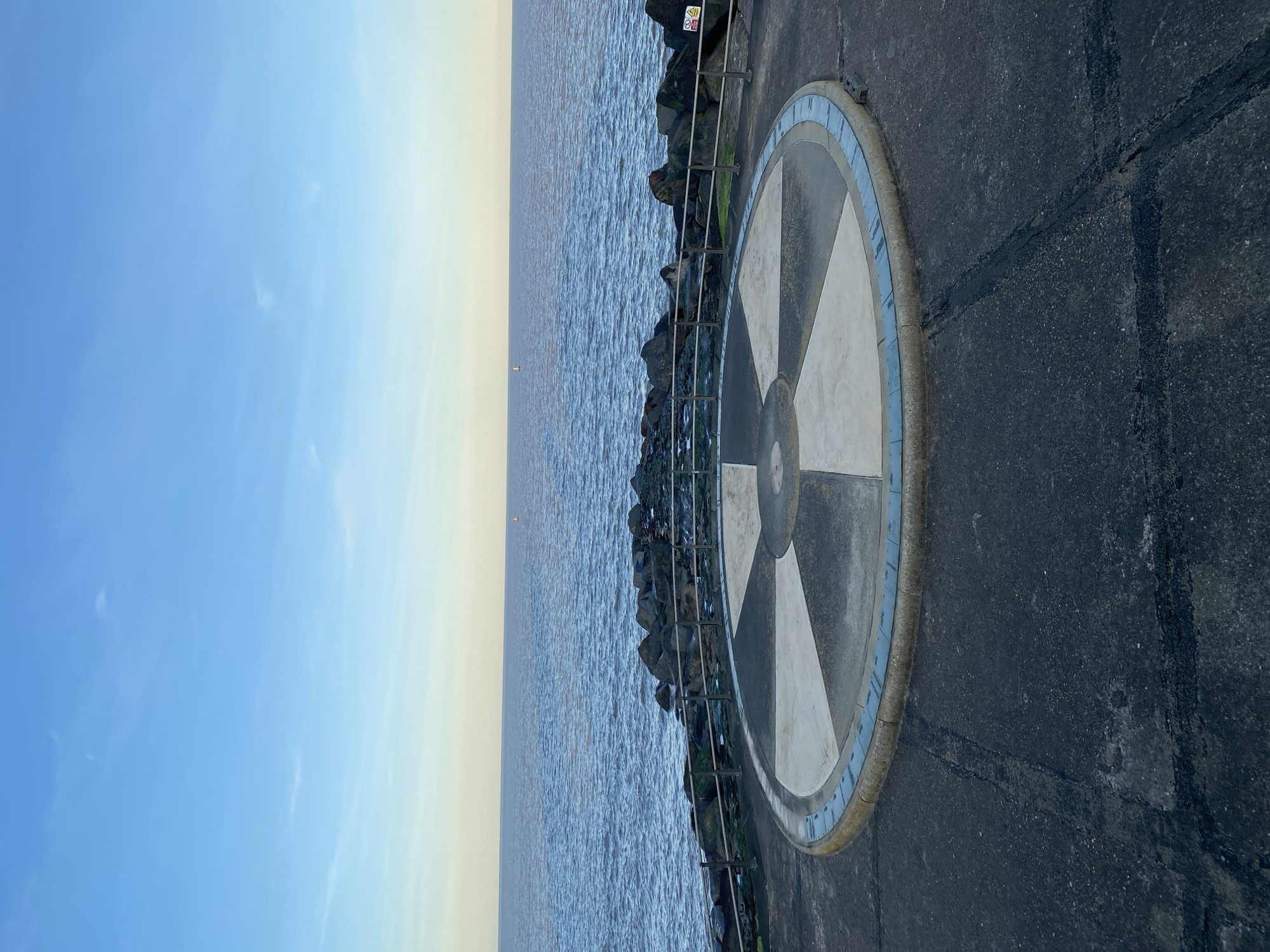 The image depicts a seaside scene in East Suffolk, United Kingdom. The foreground shows a concrete walkway featuring a circular compass rose design. To the left of the walkway, a series of rocks and a metal railing separate the walkway from the ocean. In the distance, the sky meets the water, creating a horizon line. The sky is blue with a hint of yellow, suggesting either sunrise or sunset. A warning sign is affixed to the railing. The water appears calm with slight ripples on its surface. Two small navigational buoys are visible offshore. The walkway shows signs of wear, with cracks and sealant present. The overall perspective is a wide shot taken at ground level, oriented vertically.