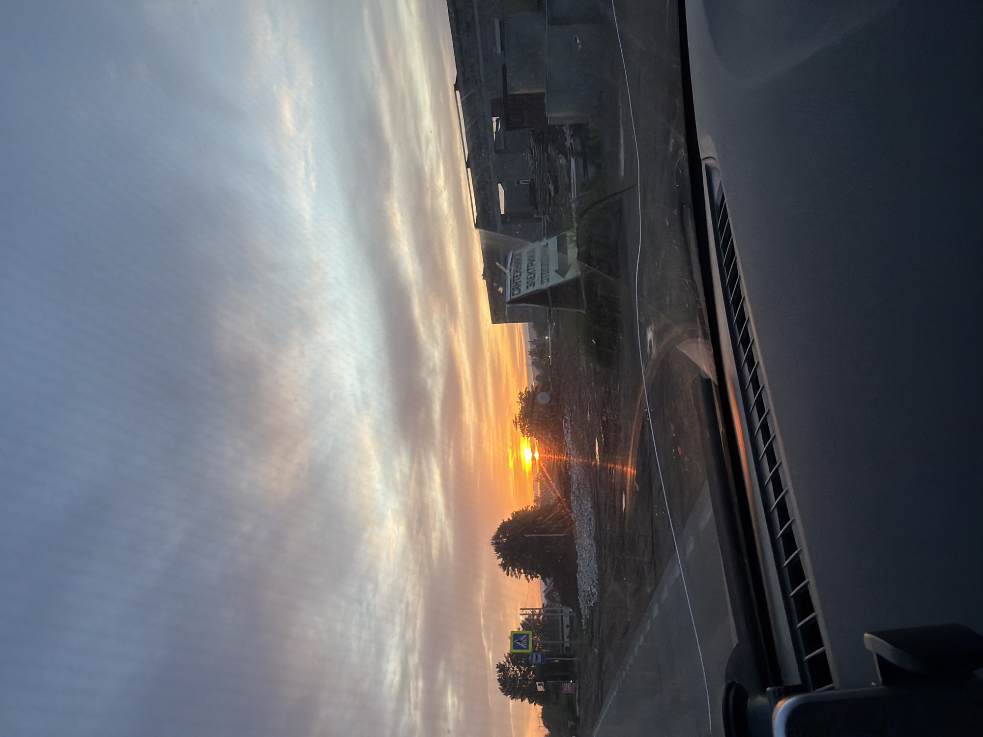 The image shows a sunset in the city of Chaikovsky, Russia, taken from inside a vehicle. The sky is filled with clouds, illuminated by the setting sun with orange hues. A building and a sign are visible to the left side of the frame. There's a road visible from the vehicle windshield.