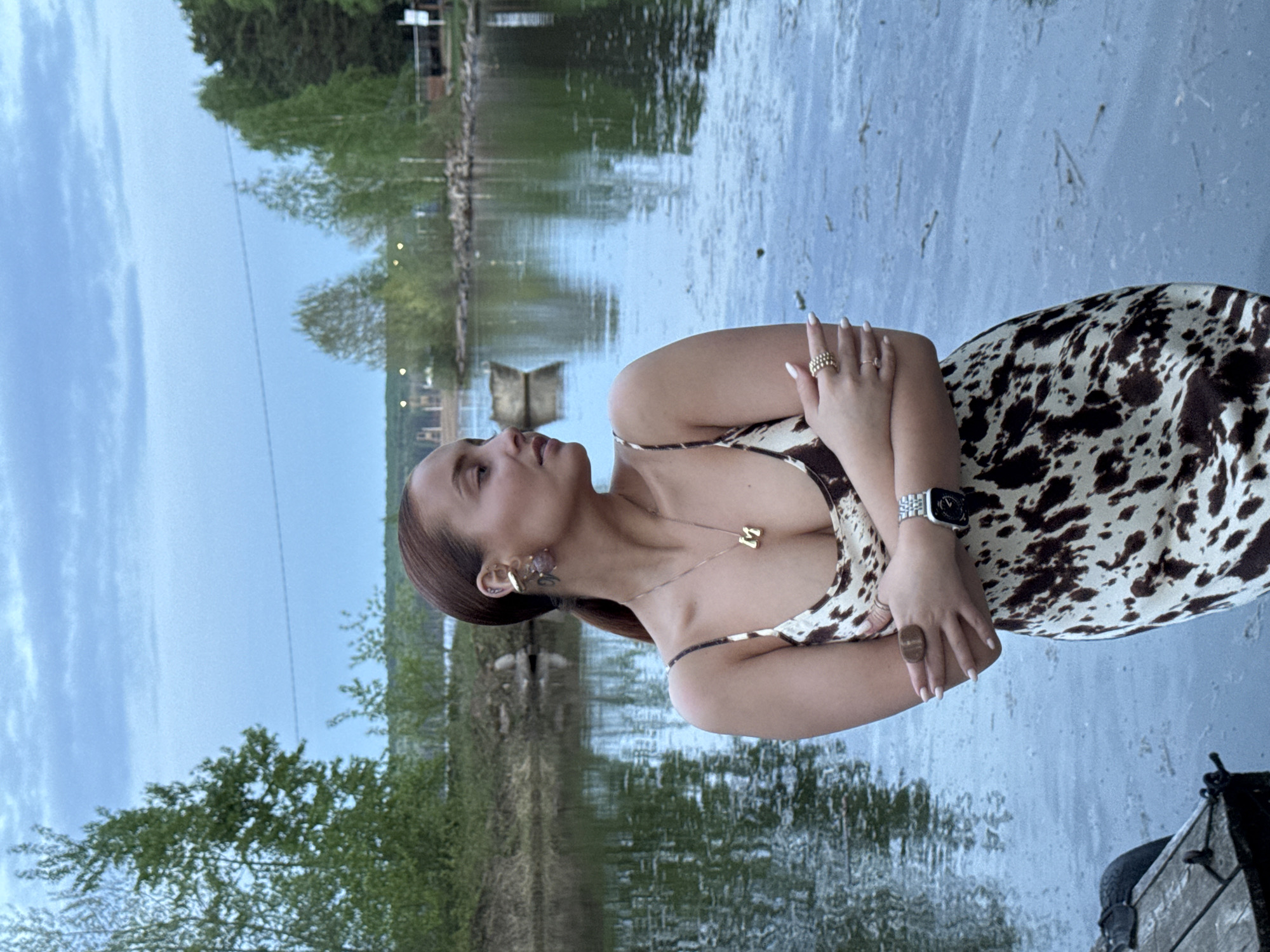 A woman in a sleeveless, brown and white cow-print dress is posing by the water. She is wearing a watch on her left wrist and multiple rings on both hands. She also has a gold necklace with a letter "M" pendant. The background shows a body of water reflecting the sky and trees. The location is in the city of Chaikovsky, Russia.