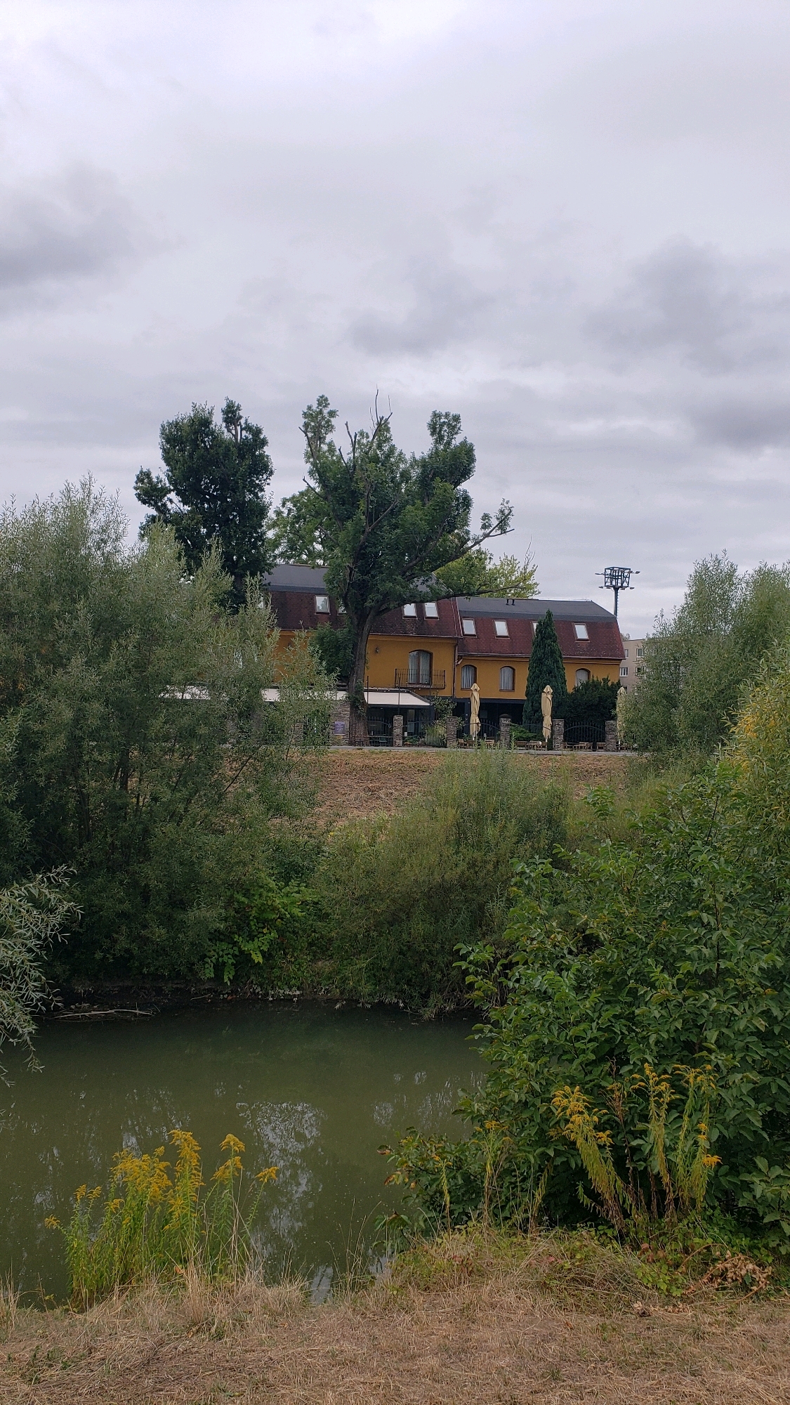 The image depicts a building with a yellow facade and a dark red roof, situated on a slope overlooking a calm body of water. The building appears to be a hotel or restaurant, with outdoor seating areas visible, including umbrellas and tables. Several trees surround the building, some of which are quite large and mature. The body of water in the foreground is bordered by dense green vegetation and dry grass on the embankment. The sky is overcast with gray clouds. In the background, a tall metal structure resembling a pylon or tower can be seen, along with other buildings further in the distance. The overall scene suggests a natural setting within or near a city. This location is identified as the area near the cable car station overlooking the city of Prešov, Slovakia, likely along the Torysa river. There are no people visible in the image.
