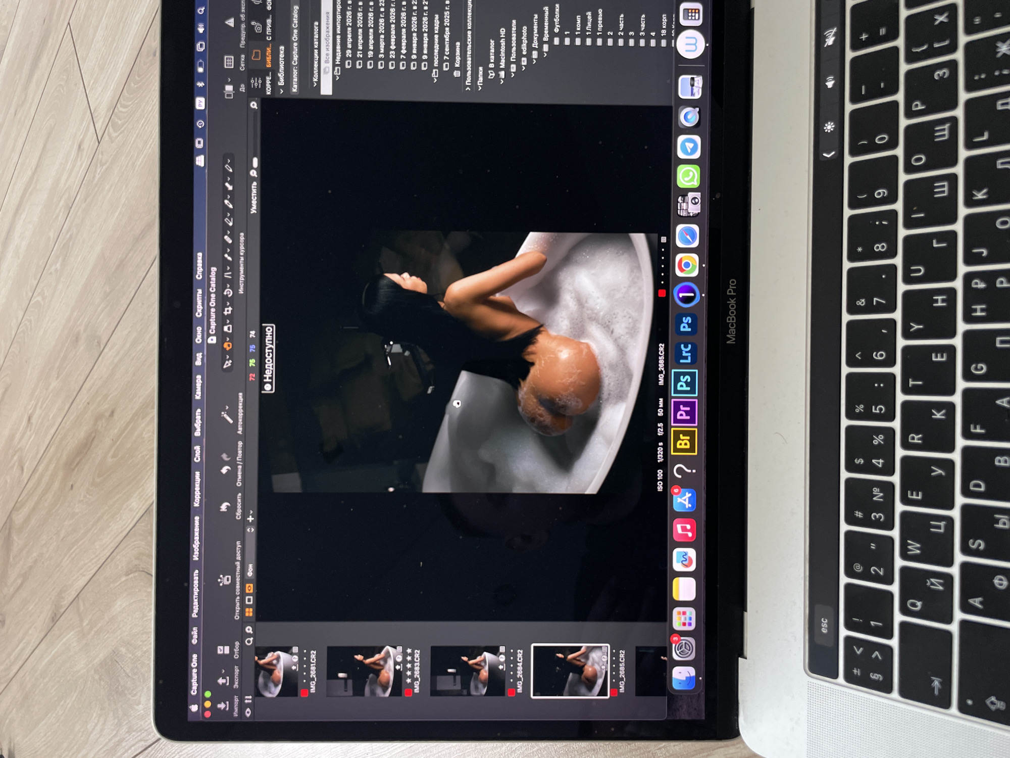 The image shows a Macbook Pro open on a wooden surface. On the screen is a photo editing program, Capture One, displaying an image of a woman in a bathtub filled with bubbles. The interface includes the menus, image thumbnails at the bottom, and editing tools. The dock at the bottom of the screen displays various applications, including a question mark, a musical note, the Safari icon, the settings icon, and others. To the right of the Macbook Pro is its keyboard with Cyrillic characters. The location is not identifiable in the image.