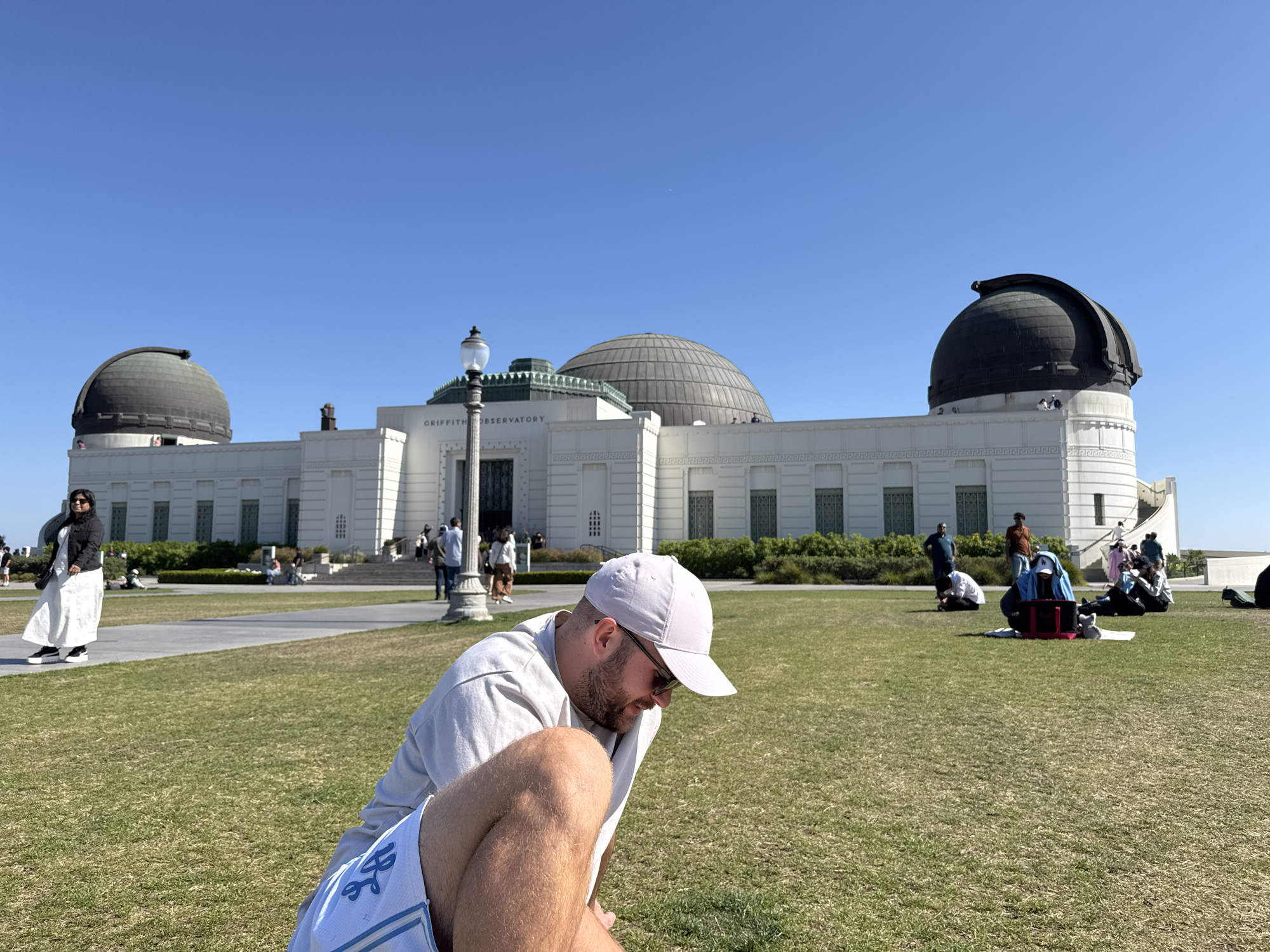 The image depicts the Griffith Observatory in Los Angeles, California, on a sunny day. The observatory's iconic white building with its domes is prominently displayed in the background. Several people are present, some walking around the observatory and others sitting or relaxing on the grassy lawn in front of it. In the foreground, there is a person sitting on the grass. The sky is clear and blue.