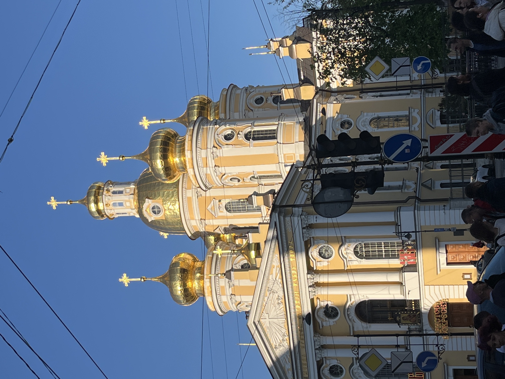 The image depicts the Smolny Cathedral in Saint Petersburg, Russia. The cathedral is a baroque-style building with a cream and white facade and five gilded domes topped with crosses. Utility wires are visible in the lower left corner of the frame, crossing the clear blue sky. A traffic light and several road signs are mounted on a pole beside the cathedral. A crowd of people can be seen in the foreground.