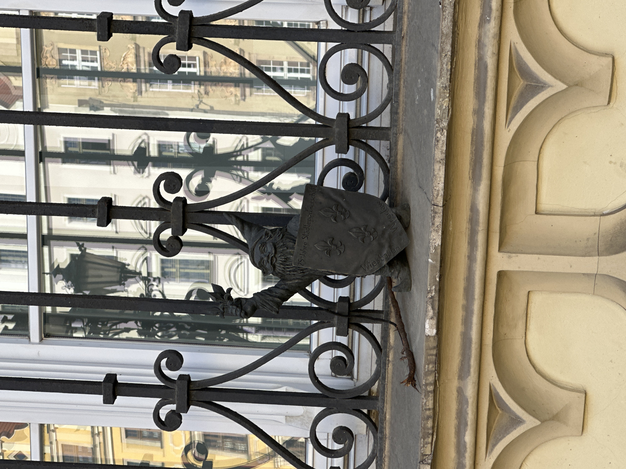 The image, rotated 90 degrees clockwise, displays a bronze gnome statue situated on a window ledge or balcony, positioned against a dark, ornate wrought iron railing. The gnome wears a pointed hat and tunic, holding a staff or scepter in its left hand and a shield in its right.
The shield is adorned with three prominent fleur-de-lis symbols. Visible text on the shield includes "25-Jahre Städtepartnerschaft" (25 Years City Partnership) and "Wiesbaden." The word "Wrocław" is also partially visible along the bottom edge of the shield.
Behind the gnome and the black wrought iron railing, large windows reflect the architectural details of other buildings, indicating an urban environment. These reflections show multiple windows and decorative elements of adjacent structures. The lighting suggests it is daytime.
To the right of the railing, part of a yellowish building facade is visible, featuring carved or molded relief patterns and details. A thin, dry branch or root extends from the base of the gnome. The wrought iron railing itself is intricately designed with scrollwork and sturdy vertical bars. The scene captures a whimsical detail characteristic of Wrocław, Poland, known for its dwarf sculptures.