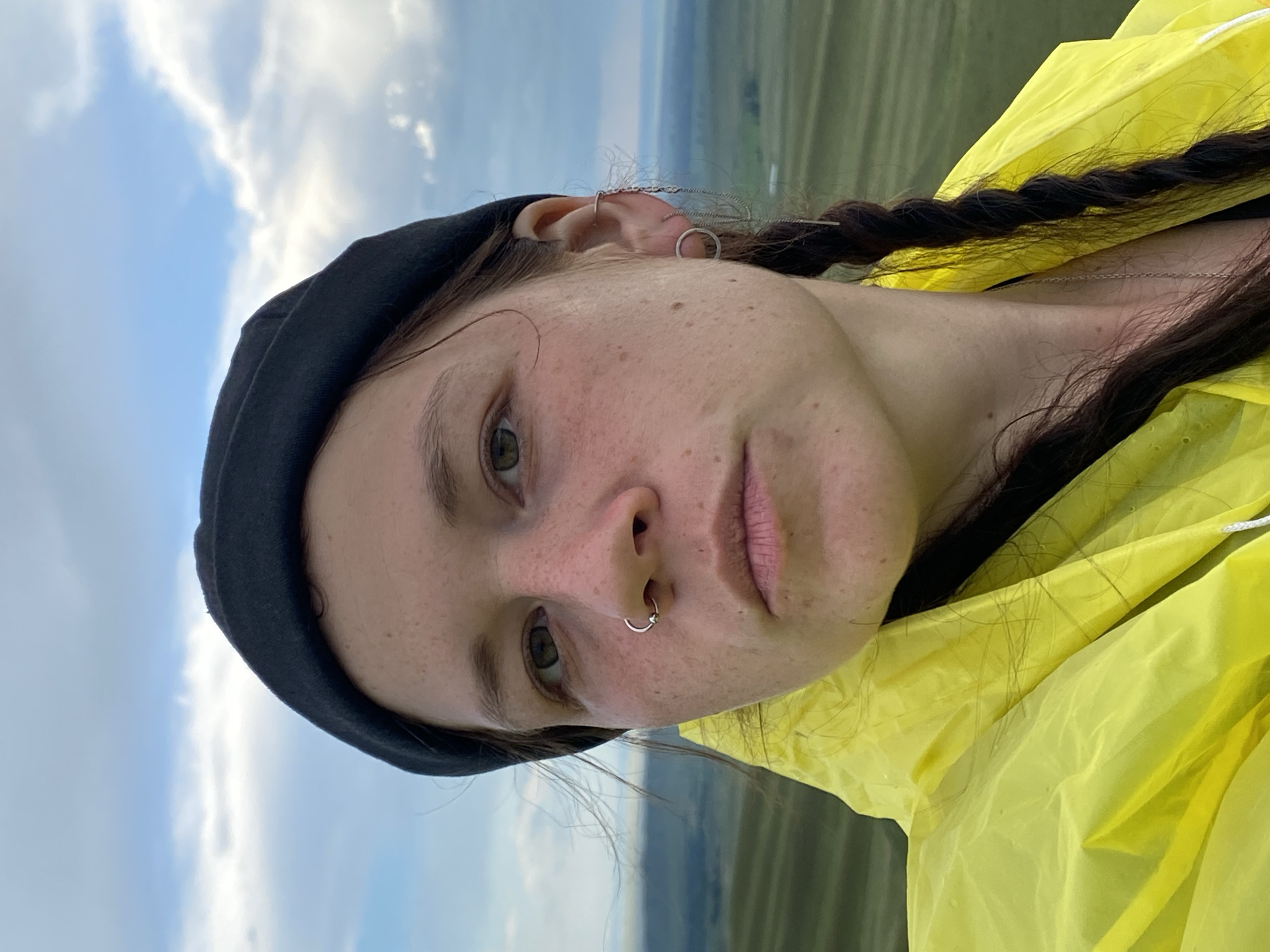 A young woman with freckles and a black beanie poses for a selfie in Verkhneozernoe, Russia. She is wearing a bright yellow raincoat. Her hair is braided and hangs over her shoulder. She has a hoop piercing in her nose and several earrings. The background shows rolling green hills under a cloudy sky.