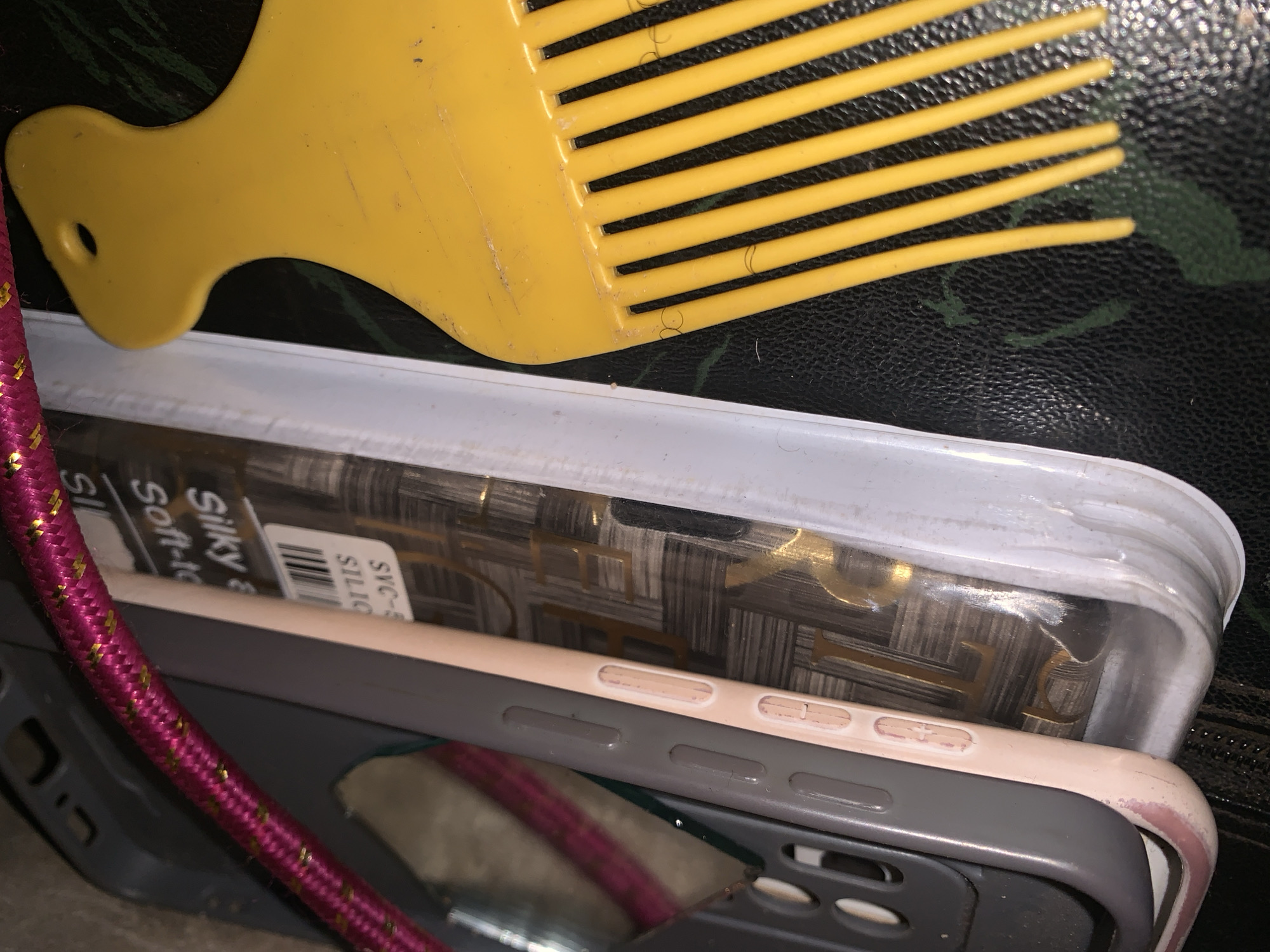 A pink and gold braided cord lies diagonally across a stack of objects on a dark, textured surface. From left to right, the objects include a gray phone case, a pale pink phone case, a clear plastic case containing a phone with a patterned back, and a bright yellow comb with wide tines. The gray phone case is partially visible on the left, showing some cutouts. The pink phone case is next, with visible buttons along its edge. Inside the clear plastic case, a phone with a repeating pattern of gold letters, possibly a designer logo, is visible. The yellow comb occupies the right side of the frame, with its tines pointing downwards. The dark surface underneath appears to be some kind of leather or textured material, with faint green markings visible. The overall impression is a collection of personal items, possibly on a table or desk, in an indoor setting.