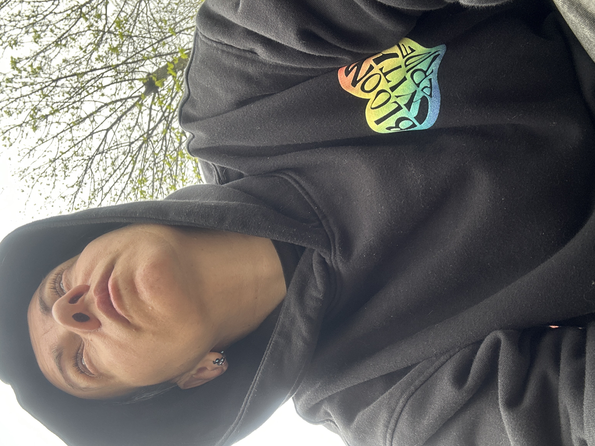 A low-angle, off-kilter photograph shows a person wearing a black hooded sweatshirt. The sweatshirt has a rainbow-colored heart-shaped emblem with the words "Bloom City Pride" on the right chest area. The person's face is visible under the hood, angled upward. They have fair skin, closed eyes, and a piercing in their ear. The background includes bare tree branches against a bright, overcast sky. The exact location in Sofia, Bulgaria, cannot be determined from the image.