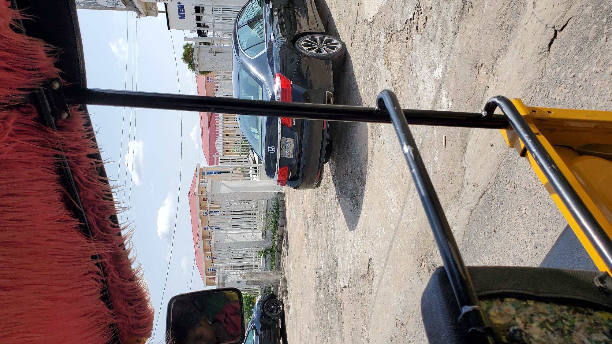 The image, taken from the perspective of a yellow motorized rickshaw, shows a street scene in Jalingo, Nigeria. A black Honda Accord sedan is parked on the right side of the frame, its rear end facing the camera. The car's license plate, RGH-769TP, is clearly visible. To the left of the car, a white fence encloses a property with buildings featuring red roofs and light-colored walls. A black car is partially visible on the far left, near a side mirror that reflects the image of a person. The ground is paved with concrete. Overhead, a bright sky with scattered clouds is visible, with the edge of a red, furry fabric canopy extending into the top of the frame.